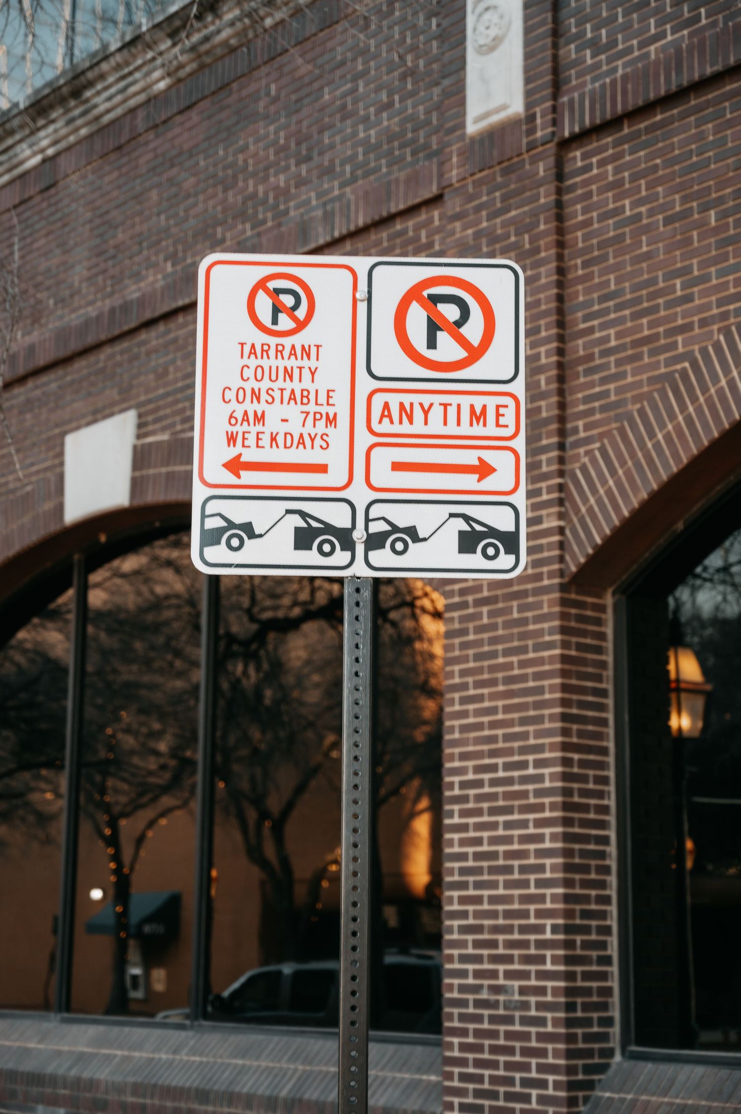No parking signs on a metal pole outside a brick building in Tarrant County, Texas, restricted 6AM-7PM weekdays.