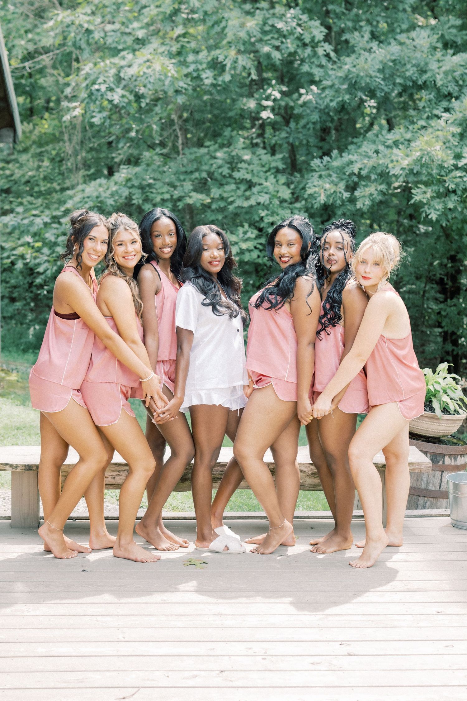 Seven bridesmaids in matching pink pajama sets pose together on a wooden deck surrounded by lush green trees.