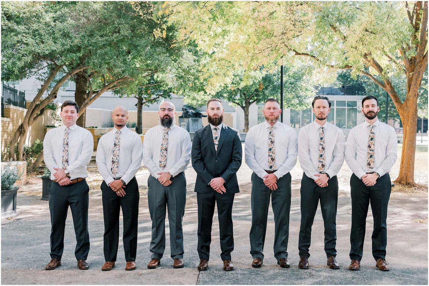 Wedding party members lined up outdoors under trees for a group photo.