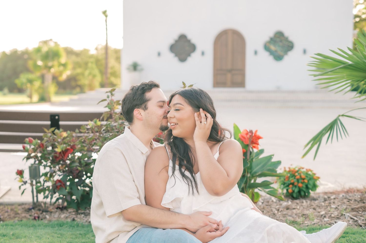 A couple shares romantic moments in a garden setting with palm trees and flowers during a sunset photo session.