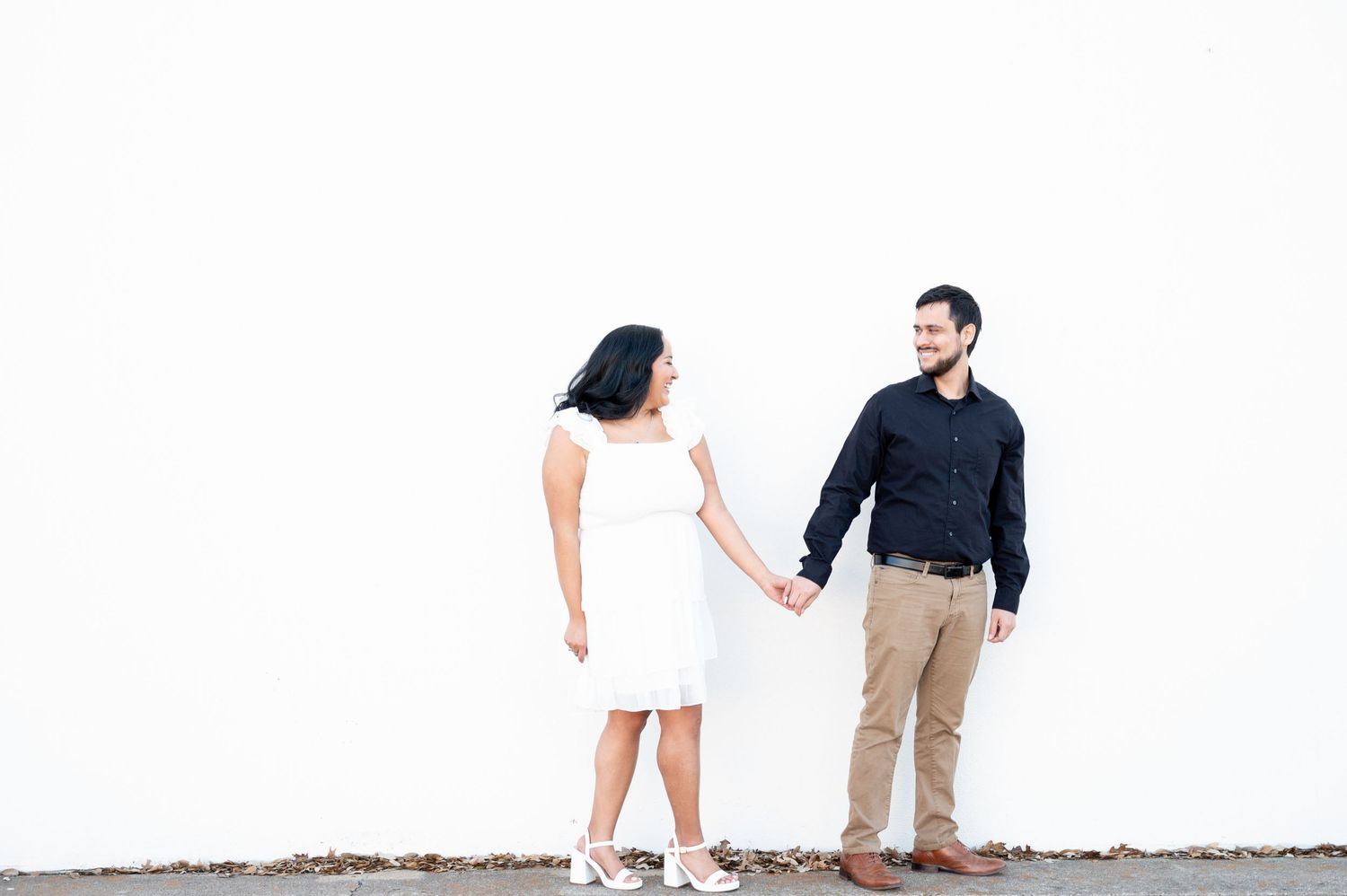Couple in contrasting outfits poses playfully against a minimalist white wall.