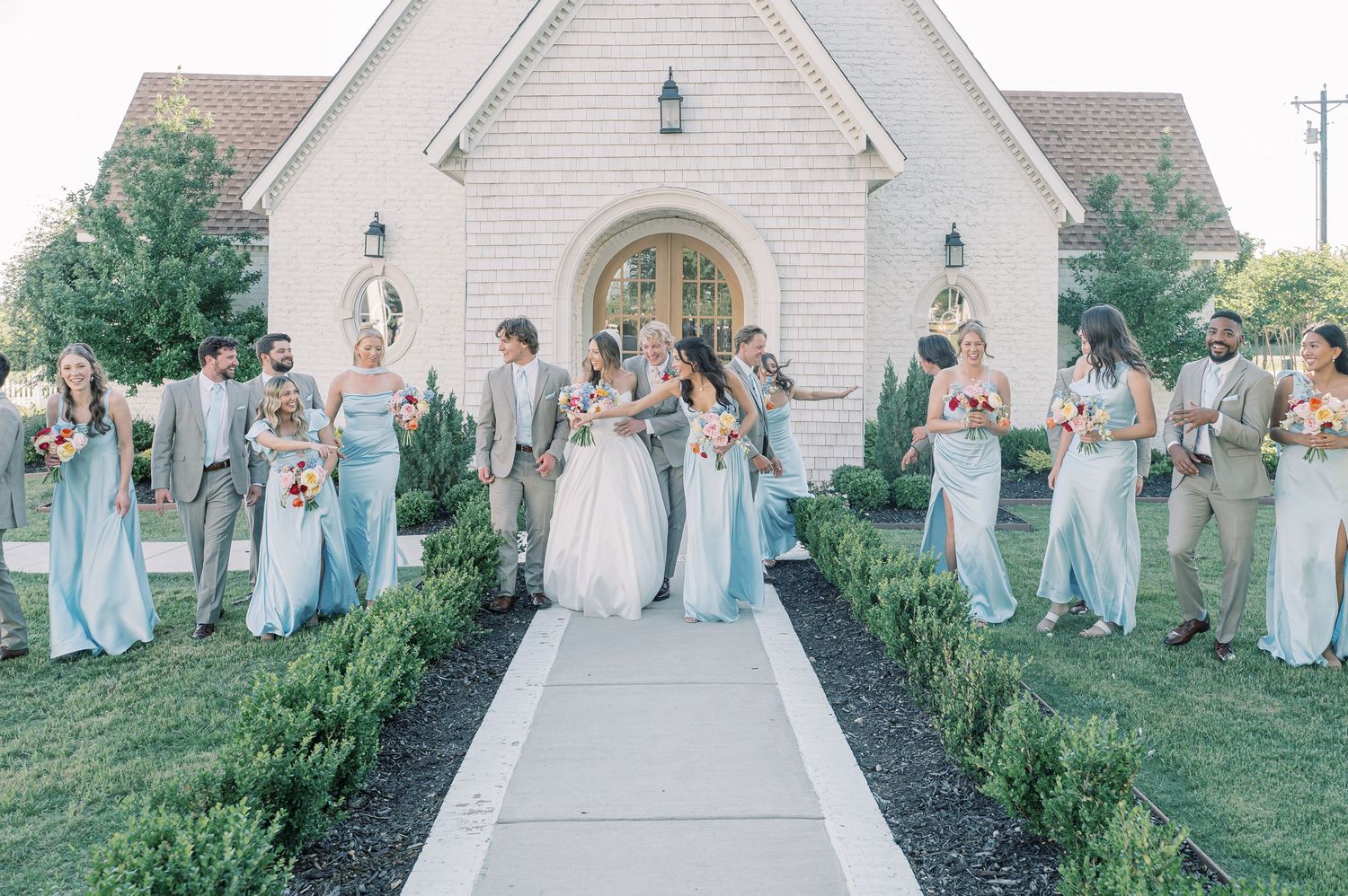Wedding party in light blue and tan attire walking outside a white chapel surrounded by lush greenery.