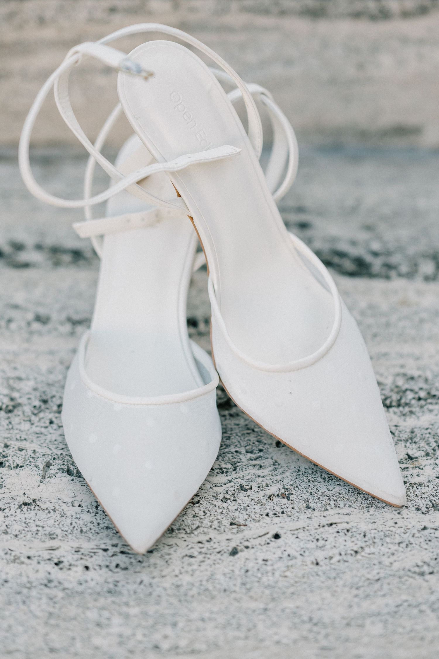 White pointed-toe bridal heels with delicate ankle straps arranged in a row on textured concrete surface.
