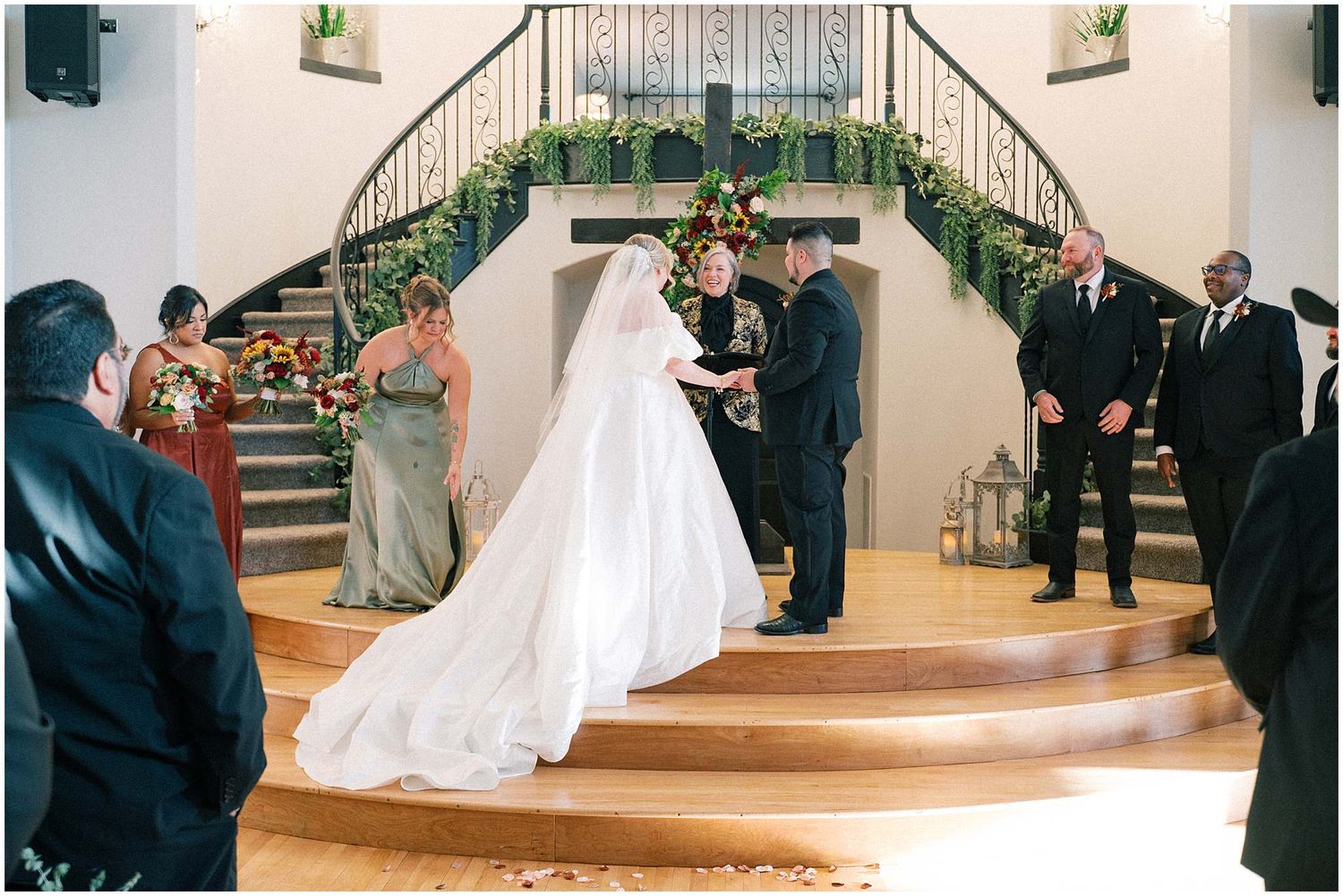 Bride and groom exchange vows on elegant curved staircase decorated with greenery during indoor ceremony.