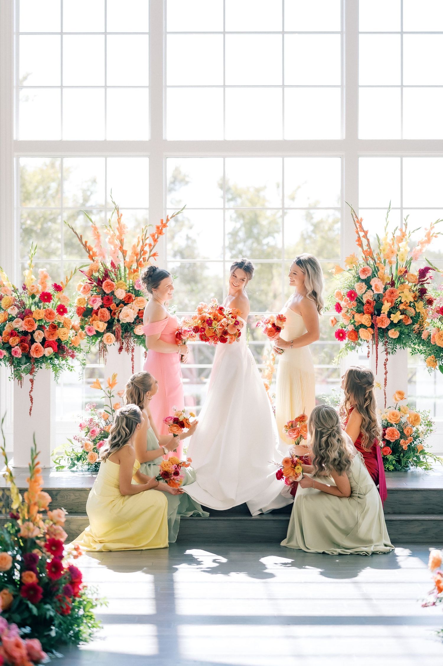 Wedding party poses in front of large windows with stunning coral and pink floral arrangements.