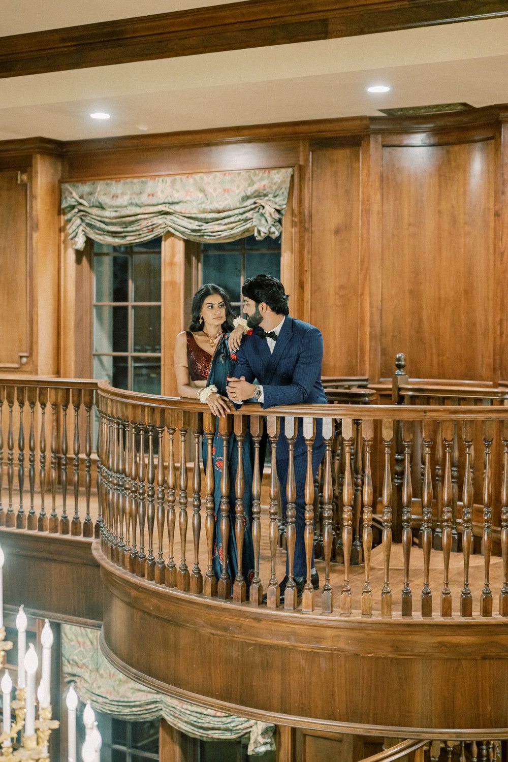 Elegant couple poses on curved wooden balcony with chandelier below.