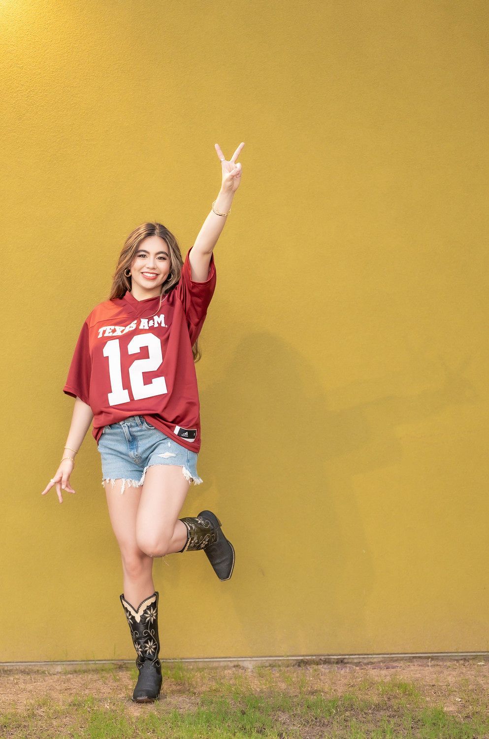 Young woman in Texas A&M #12 jersey and denim shorts making peace sign against yellow wall.
