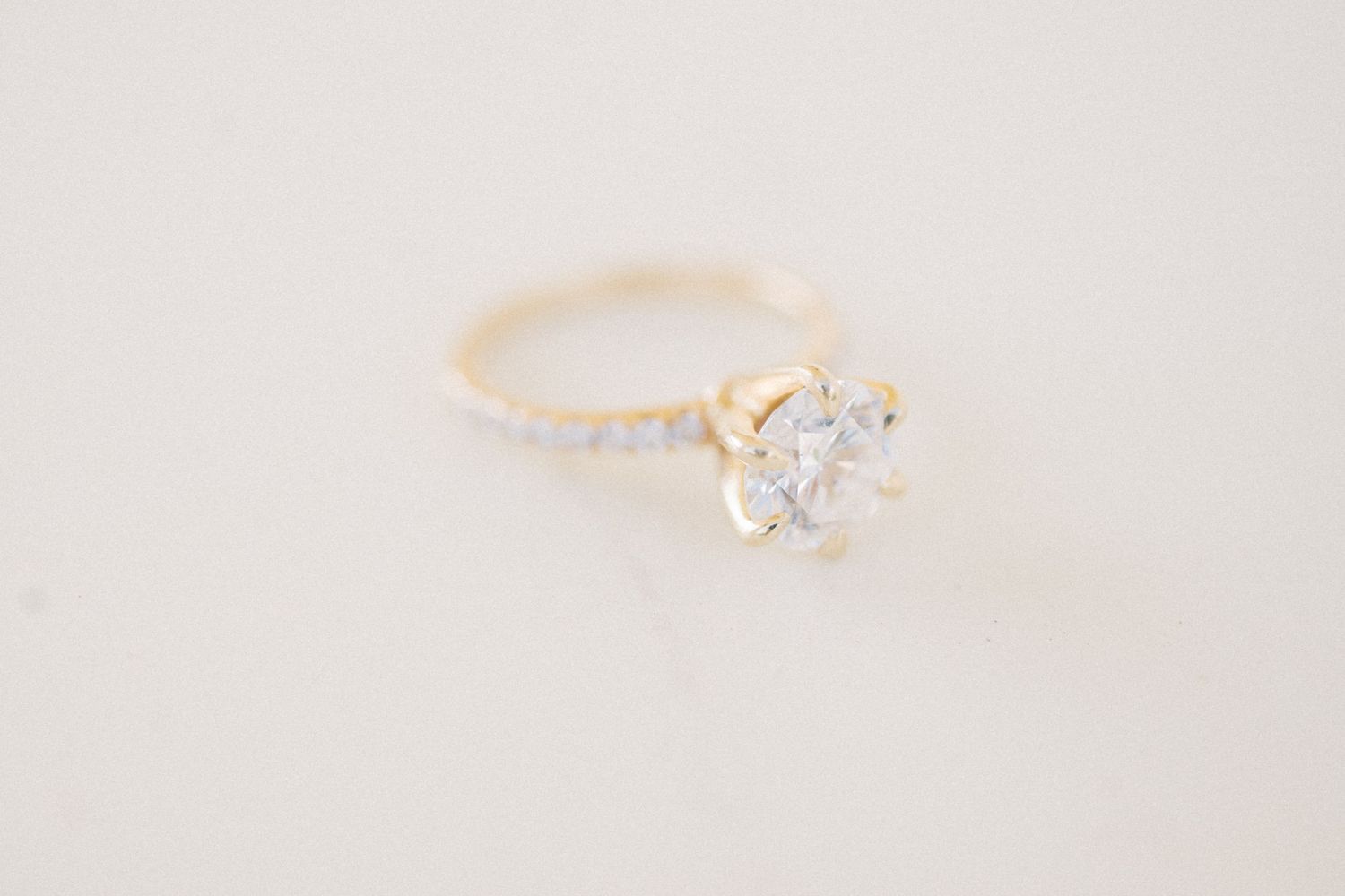 Gold solitaire engagement ring with round diamond center stone and pavé diamond band on white background.