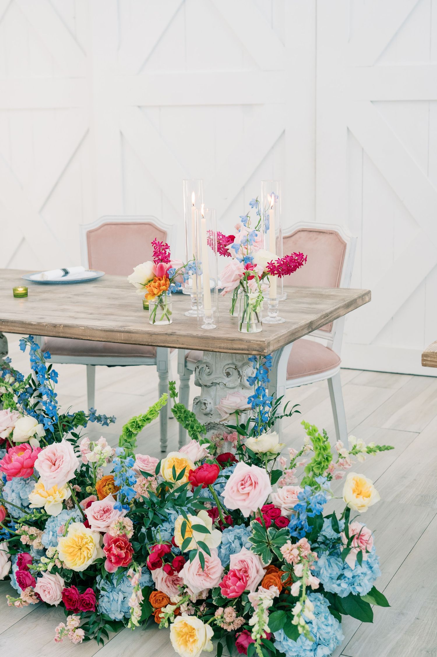 Elegant wedding table with colorful floral arrangement, pink chairs, candles, and vibrant blooms on white background.