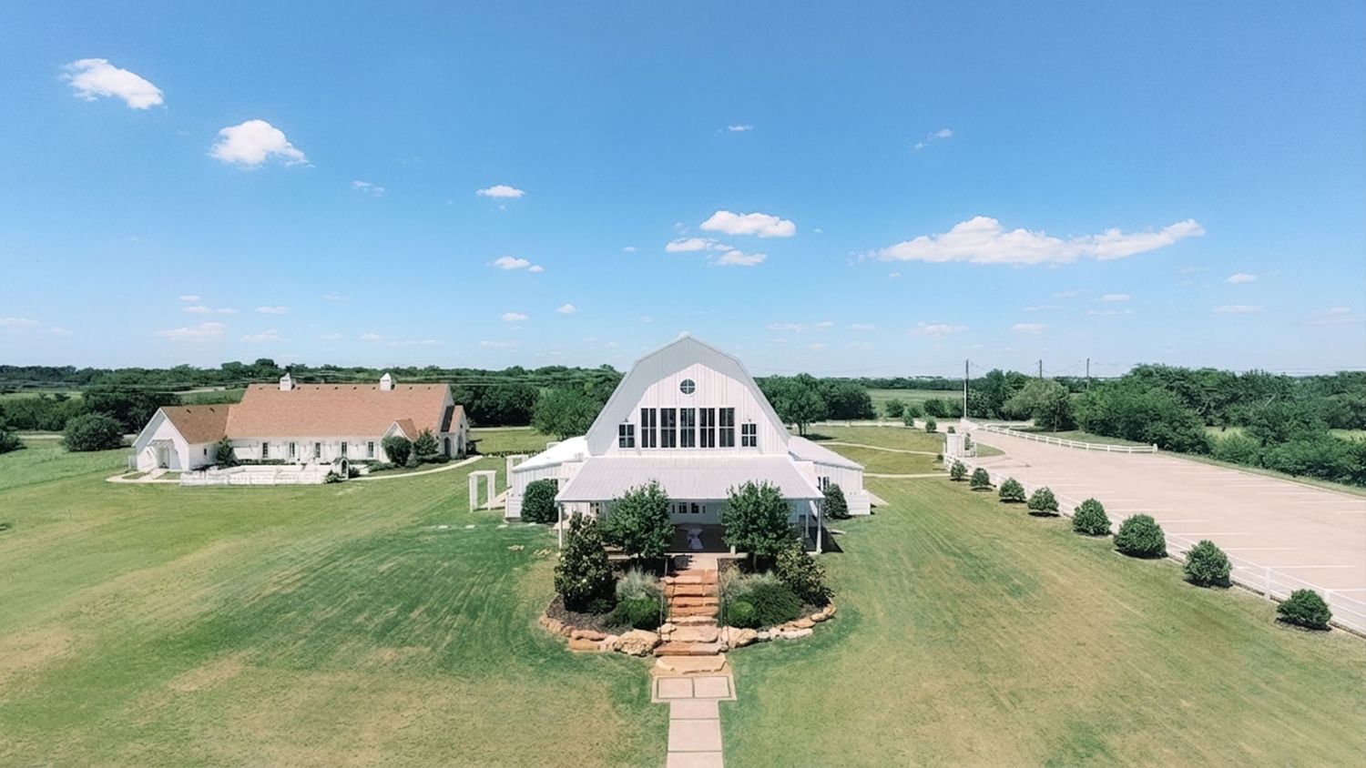 Aerial view of a white barn wedding venue surrounded by lush green lawns and manicured landscaping under a bright blue sky.