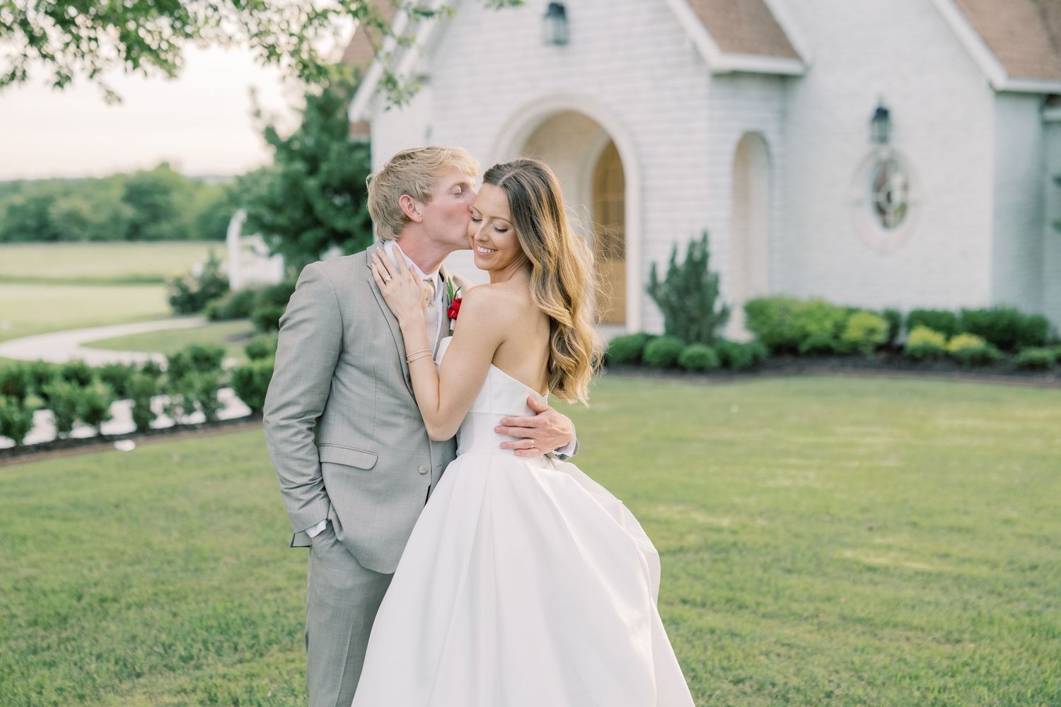 Bride and groom sharing a romantic embrace on green lawn before a charming white chapel at a summer wedding.
