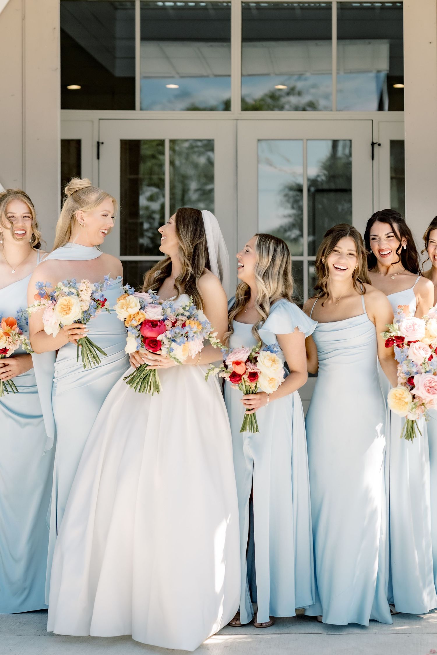 Bride in white gown with veil laughing alongside bridesmaids in light blue dresses holding colorful bouquets.