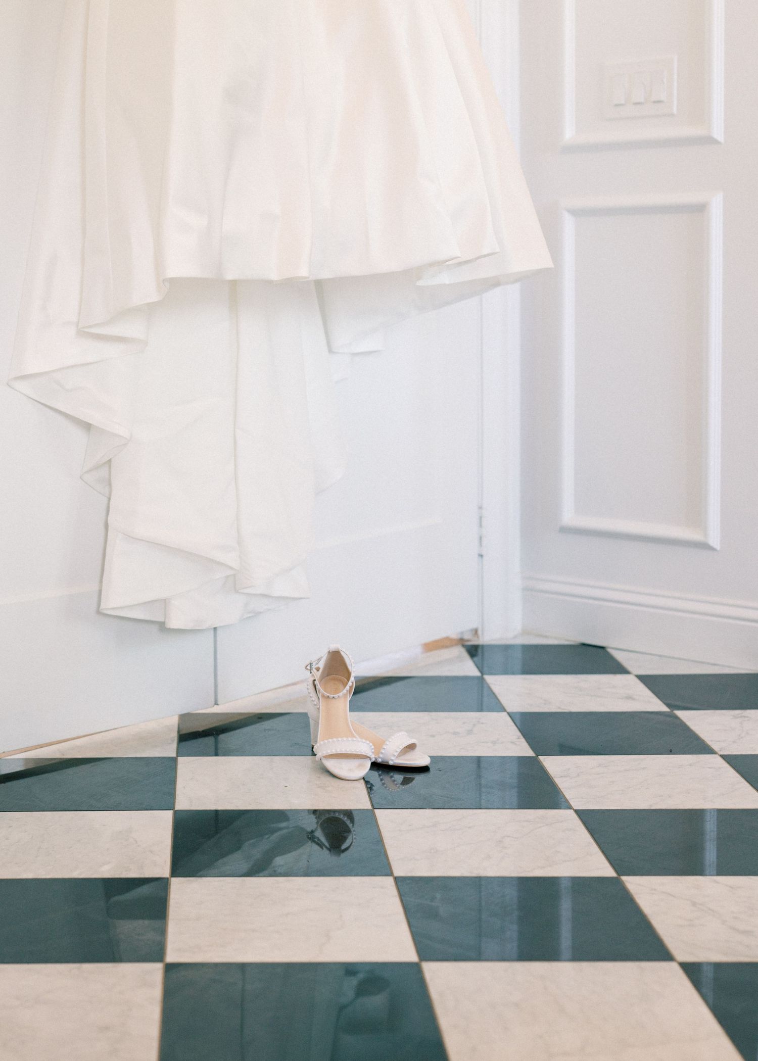 White bridal gown hanging above white strappy heels on elegant green and white checkered marble floor.