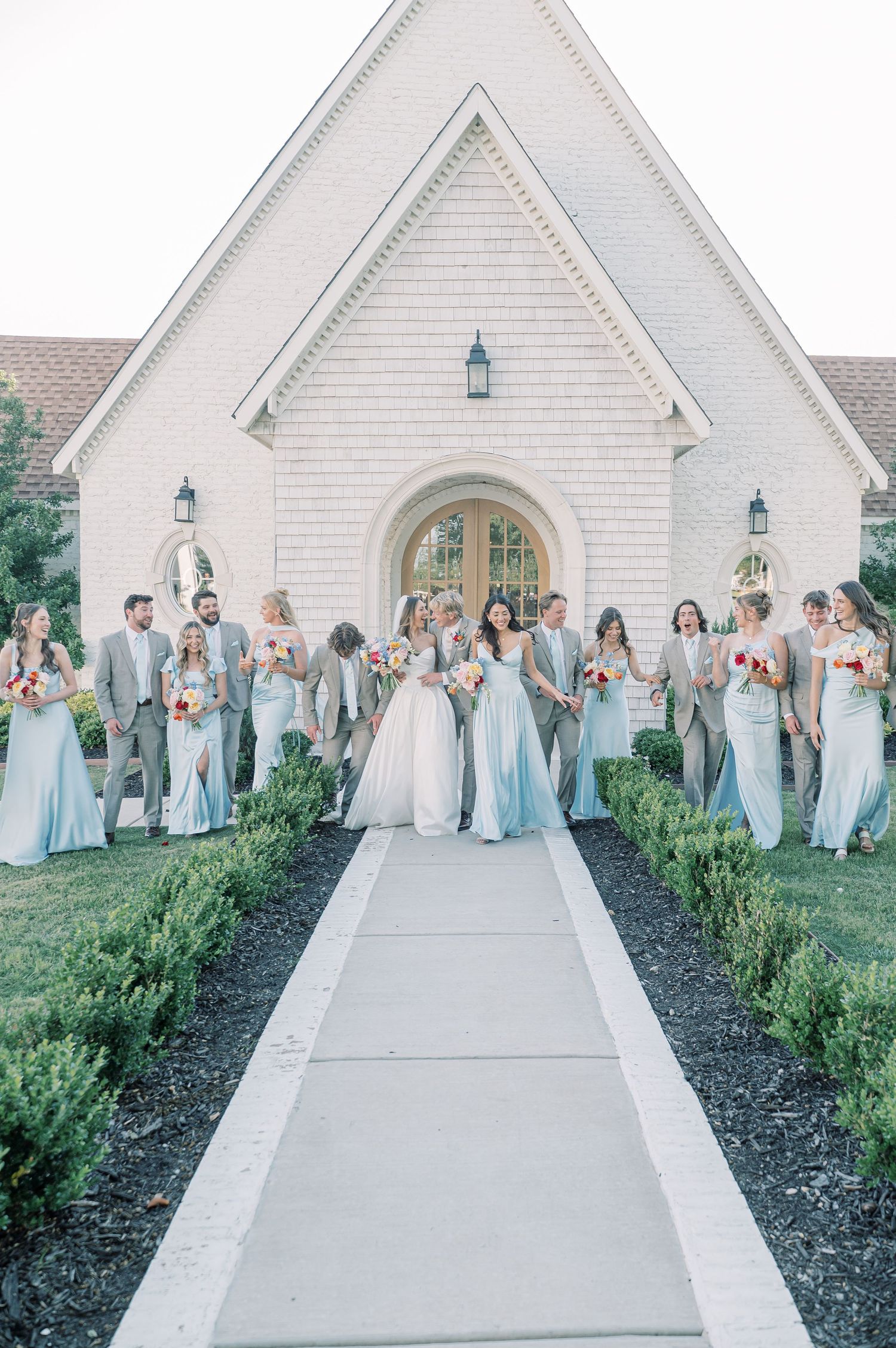 Wedding party in light blue gowns and gray suits poses on chapel pathway with bride and groom centered.