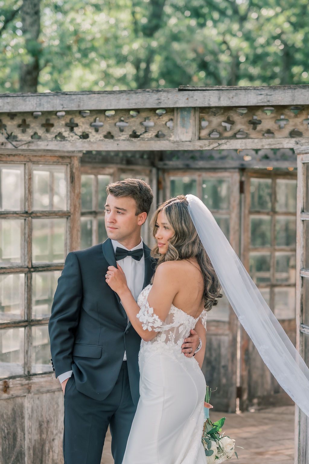 Bride in backless lace gown with veil embraces groom in navy tuxedo before rustic greenhouse backdrop.