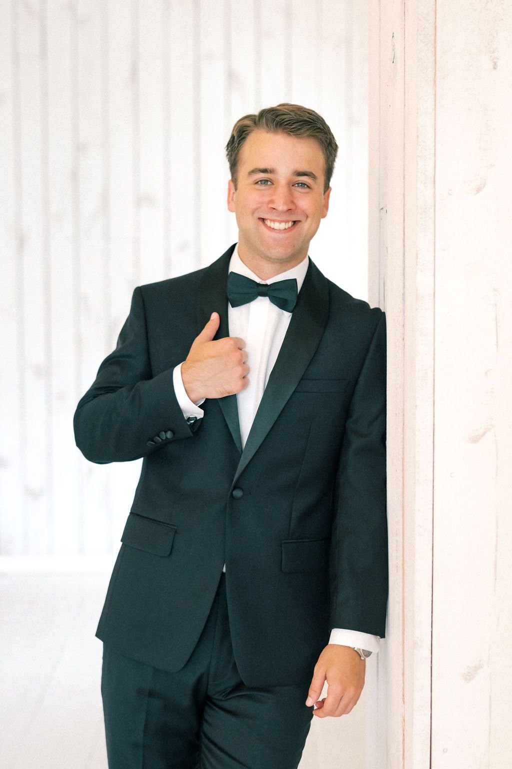 Smiling groom in black tuxedo and bow tie giving thumbs up while leaning against white wooden wall.