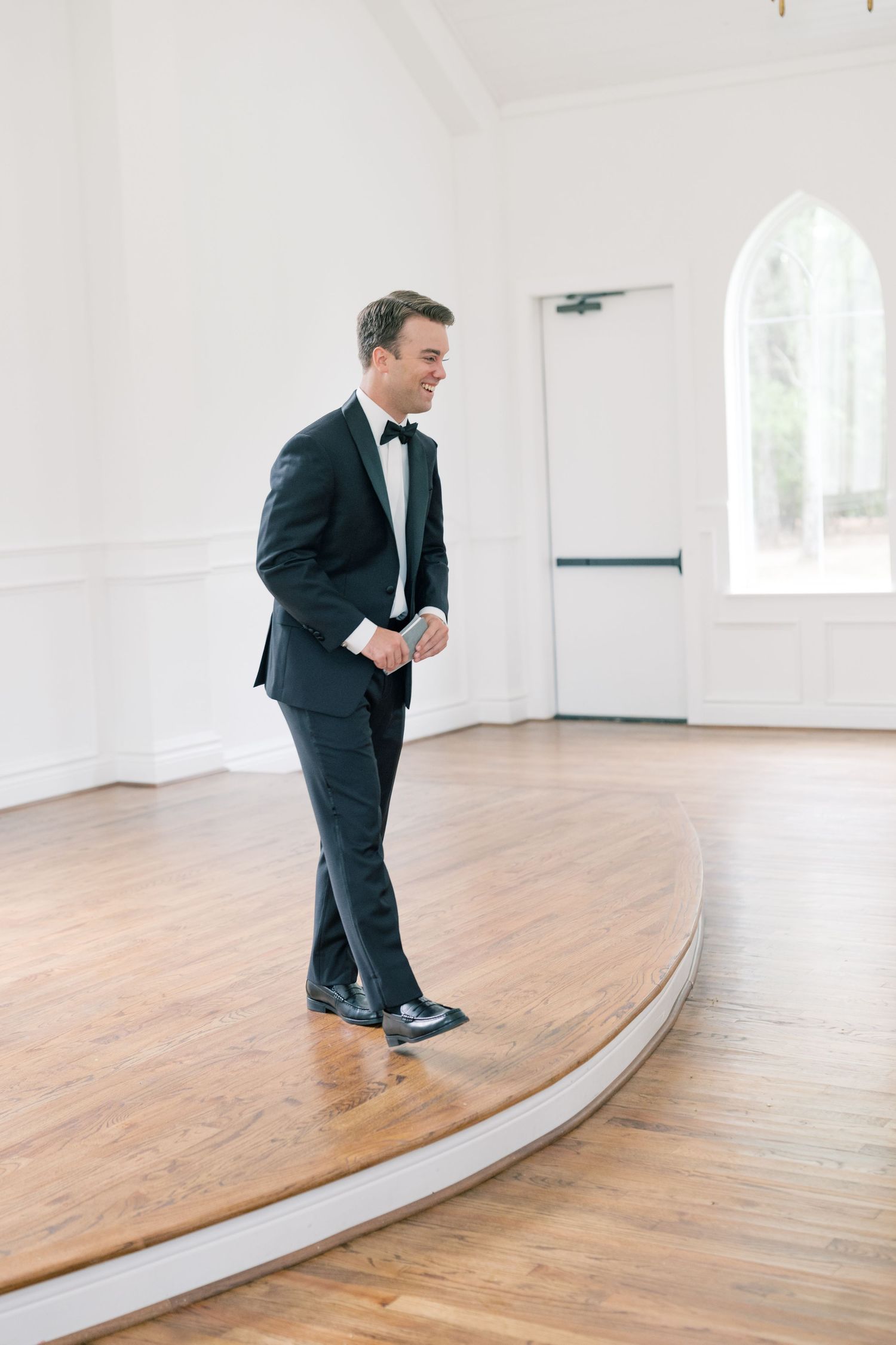 Groom in classic black tuxedo with bow tie standing in bright white chapel with arched window.
