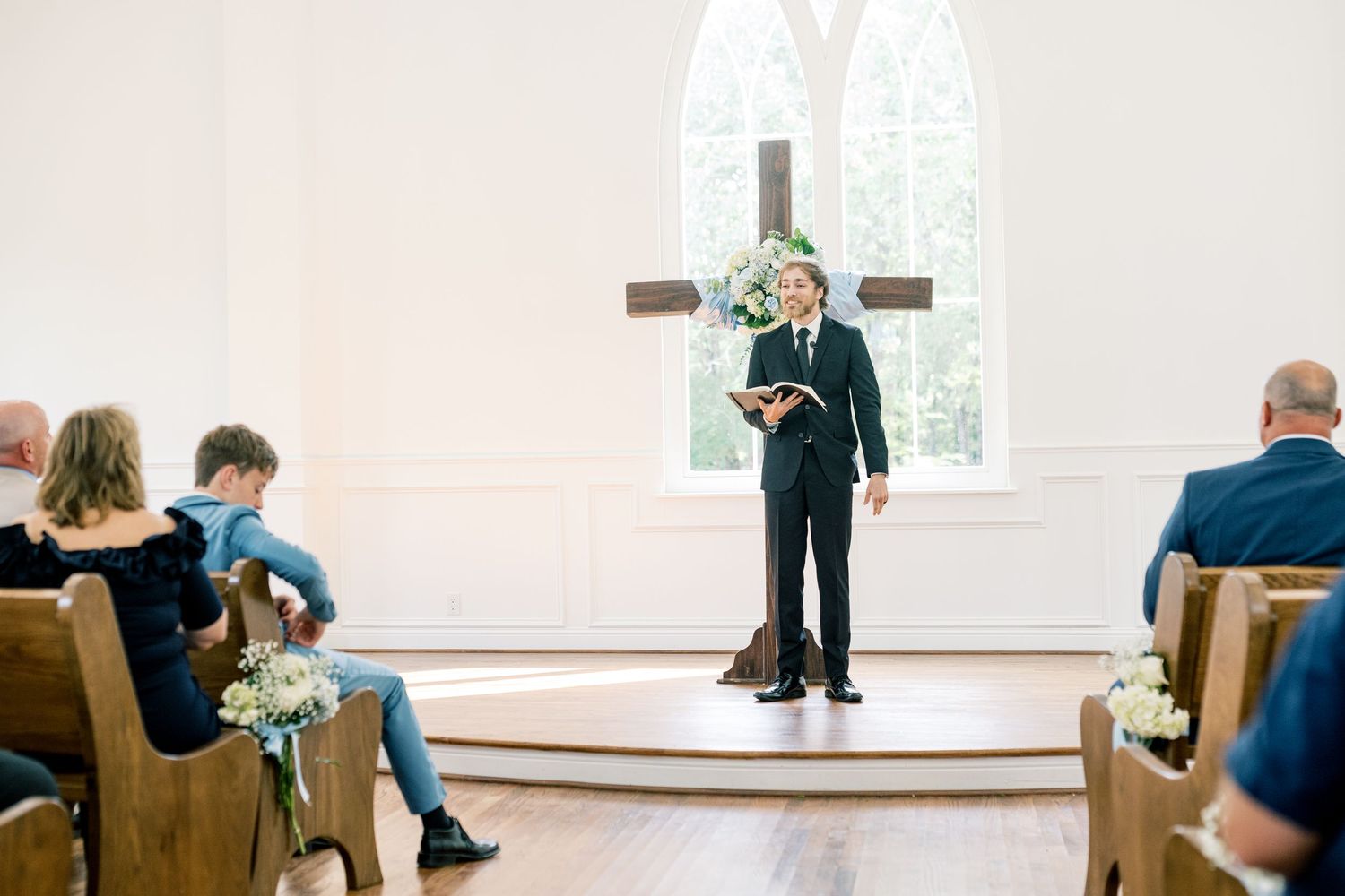 Officiant stands at chapel altar holding book, addressing seated wedding guests in a bright white church.