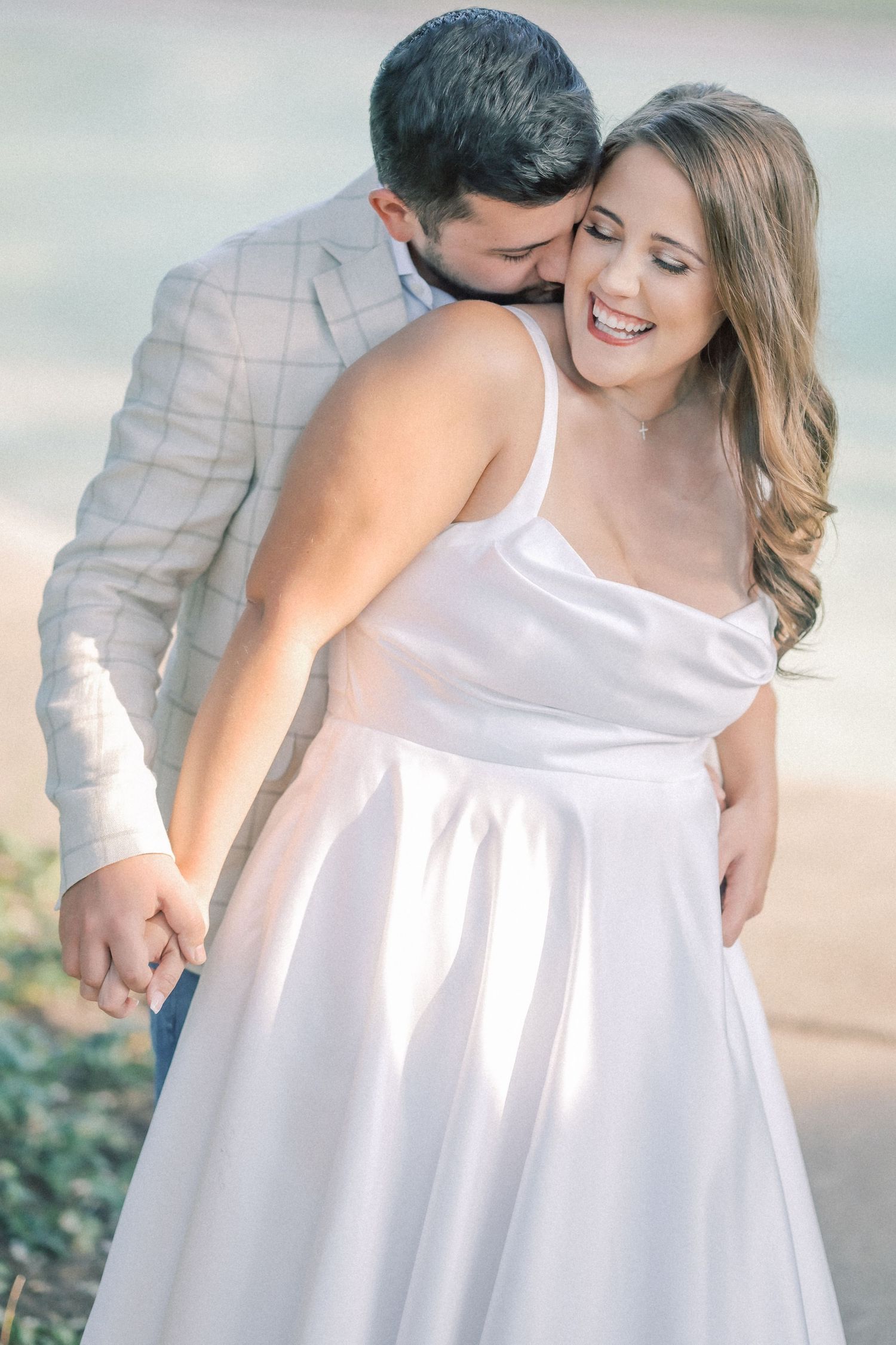 Man kissing a laughing woman on the cheek from behind as she wears a white satin dress in a soft-lit engagement photo.