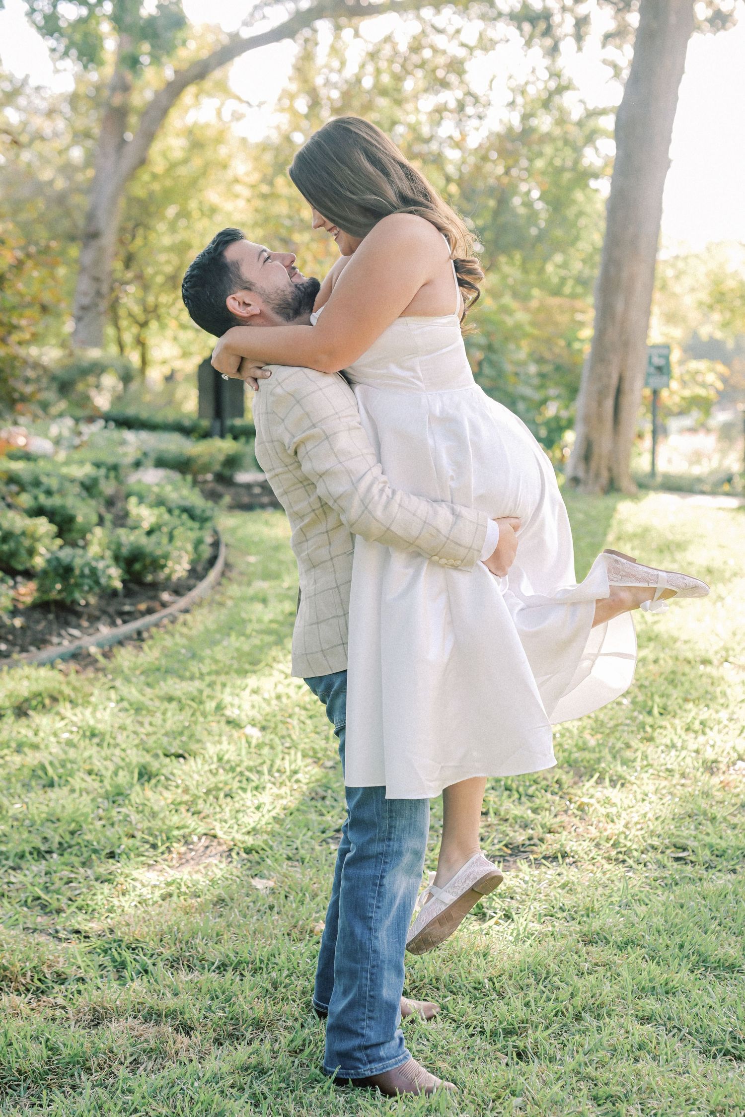Man lifts his laughing fiancée in a white dress off the ground on a grassy lawn in a park during a joyful engagement photo shoot.