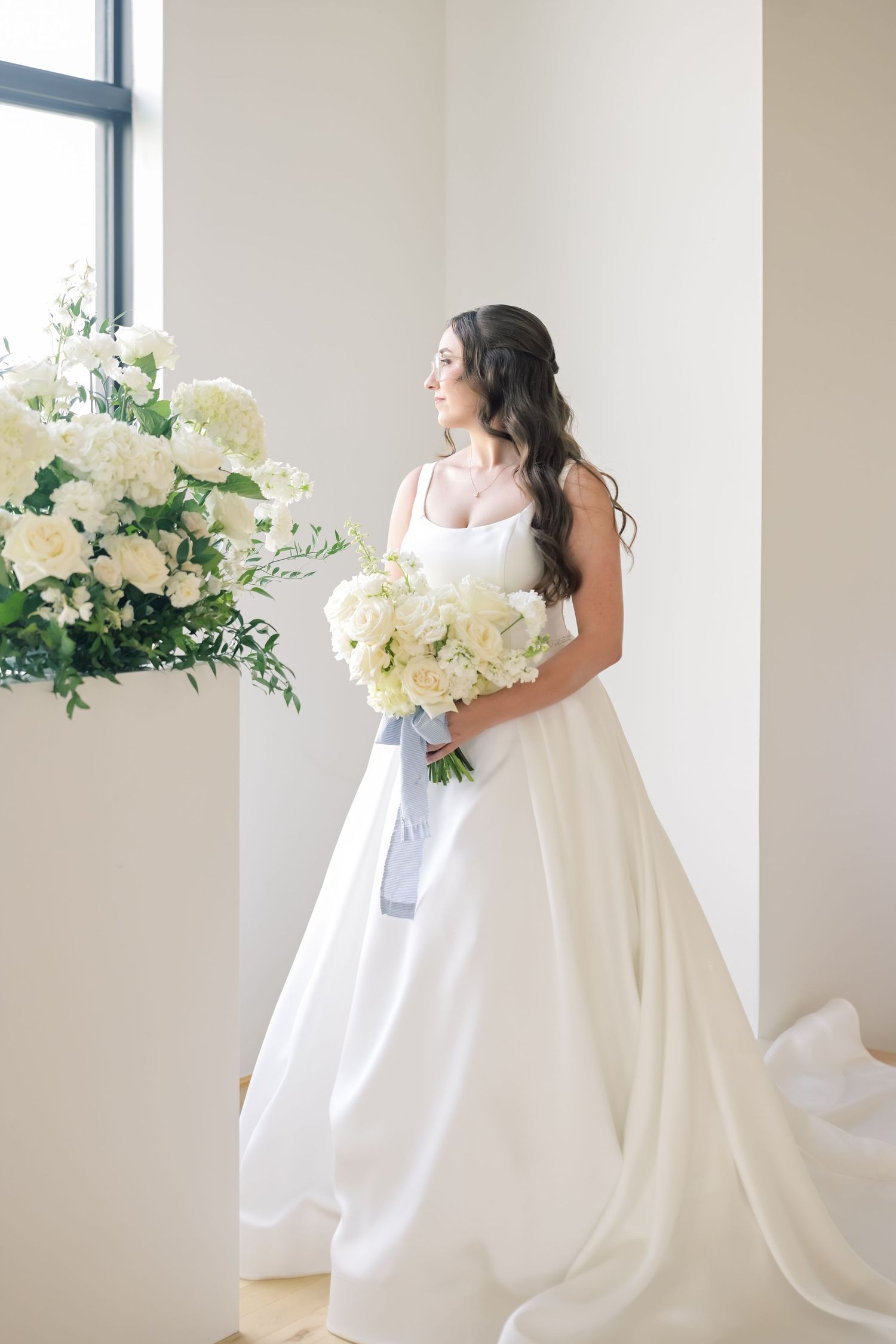 Elegant bride in white satin ball gown holds white floral bouquet beside large flower arrangement in bright room.