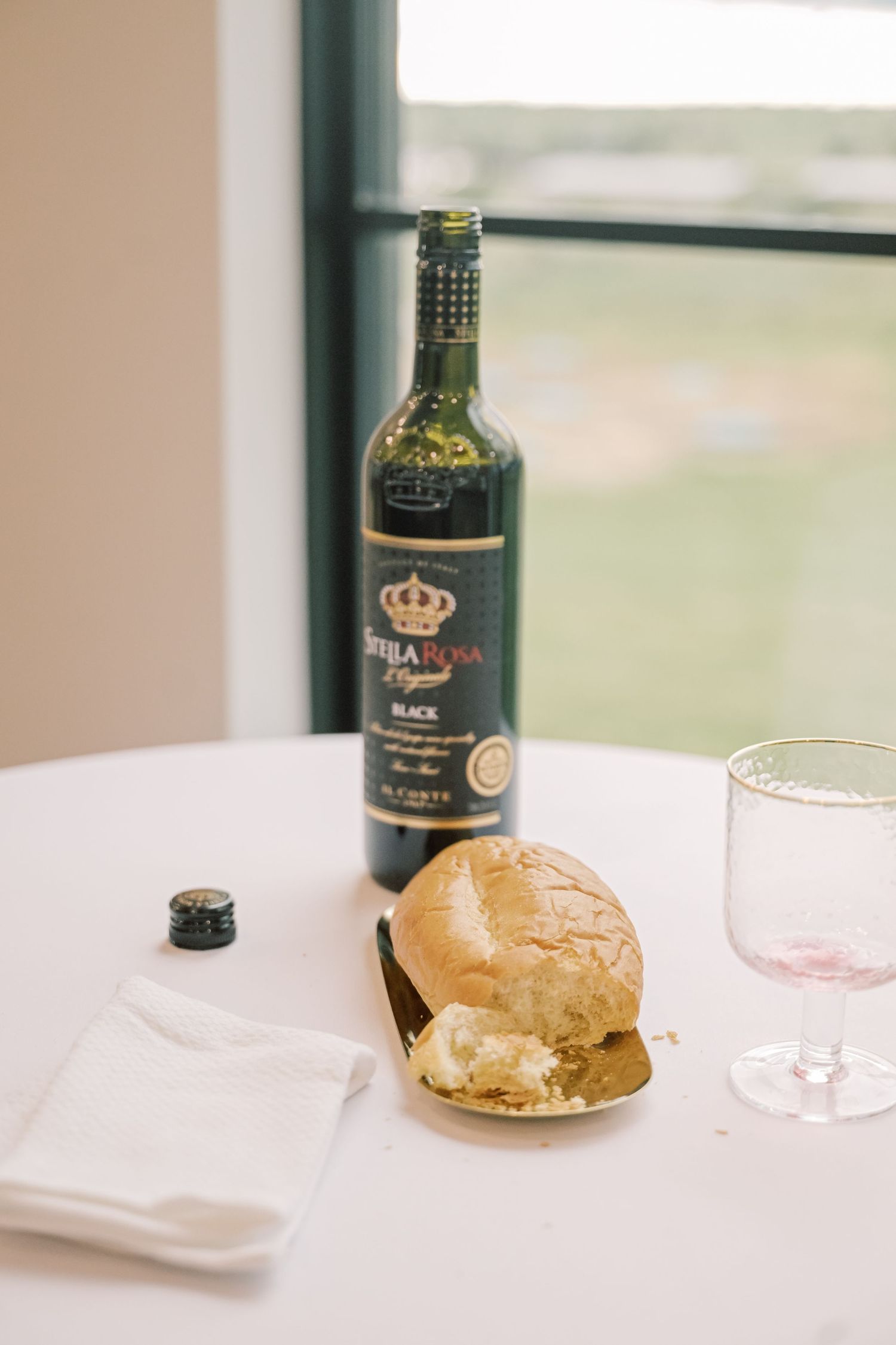 Wine bottle and bread roll on a white tablecloth beside a pink glass at a wedding reception table.