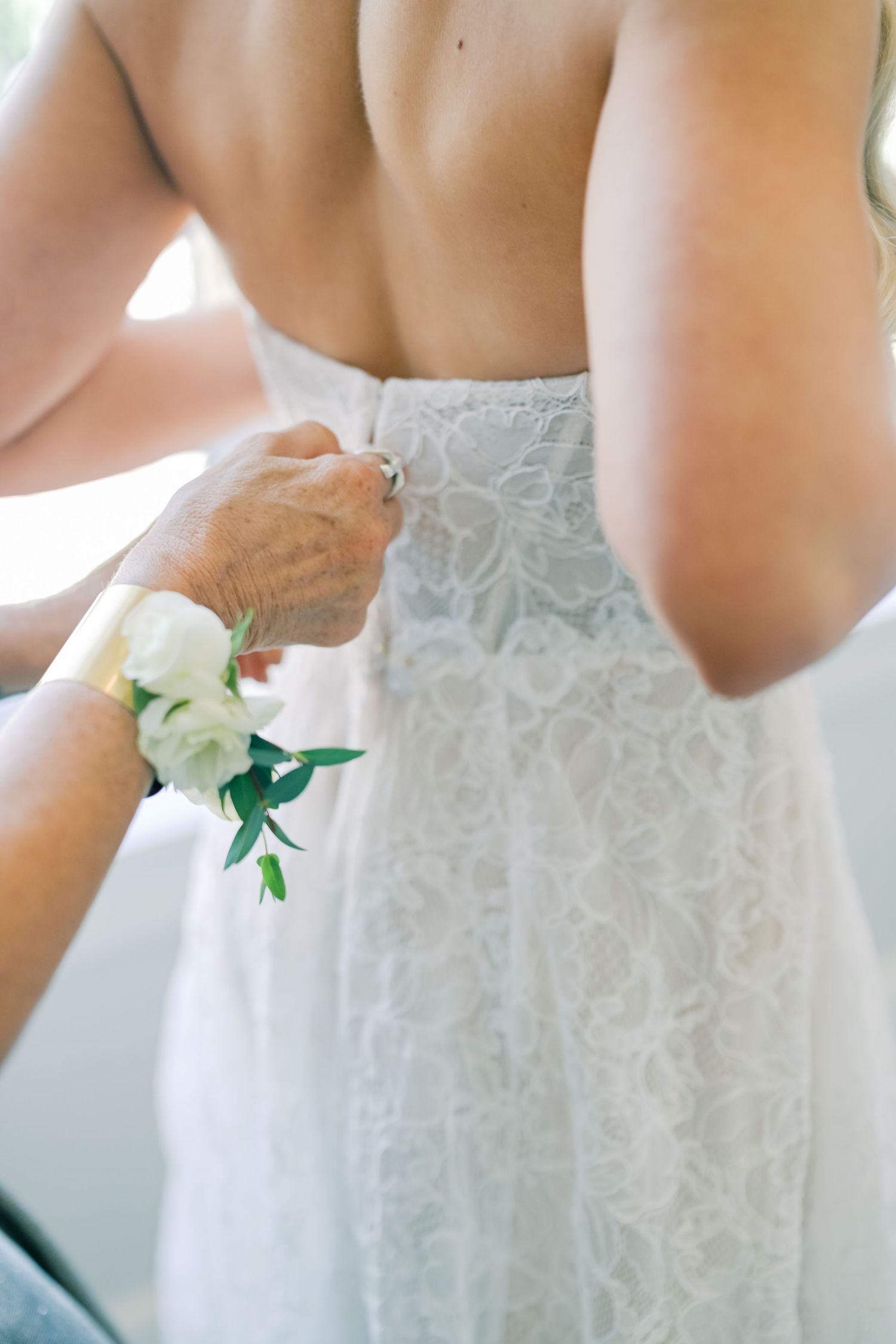 Mother fastening buttons on back of bride's white lace wedding dress while wearing floral wristlet.