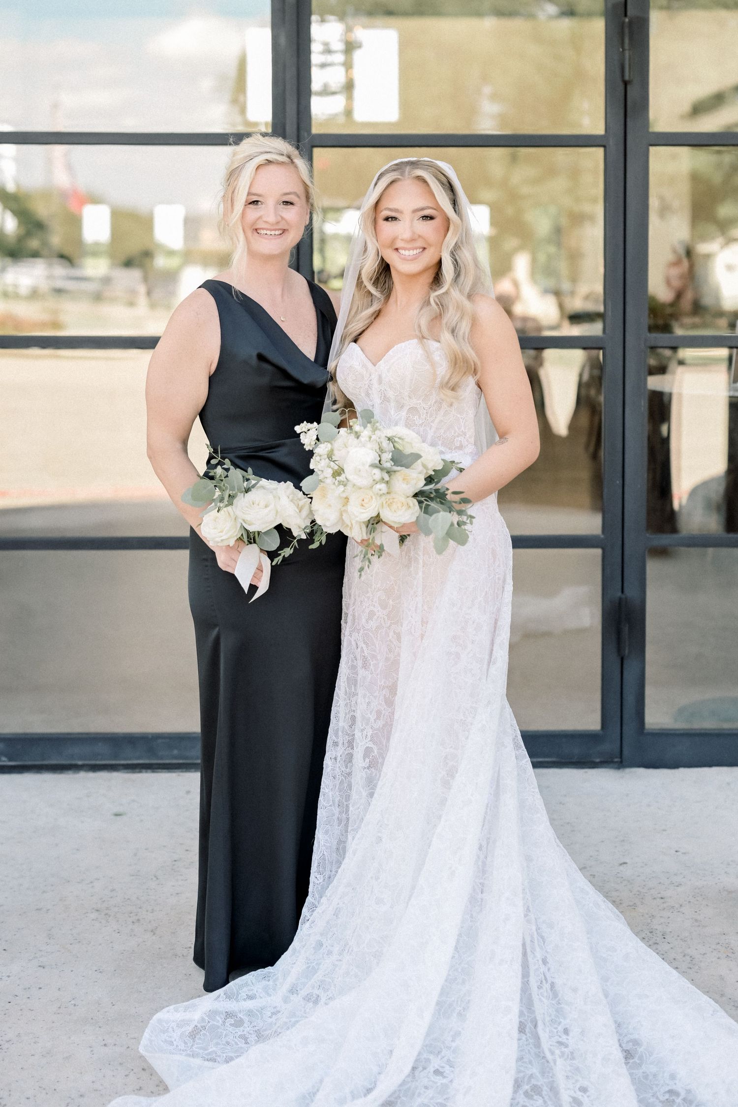 Bride in lace gown and woman in black dress hold white floral bouquets outside glass doors.