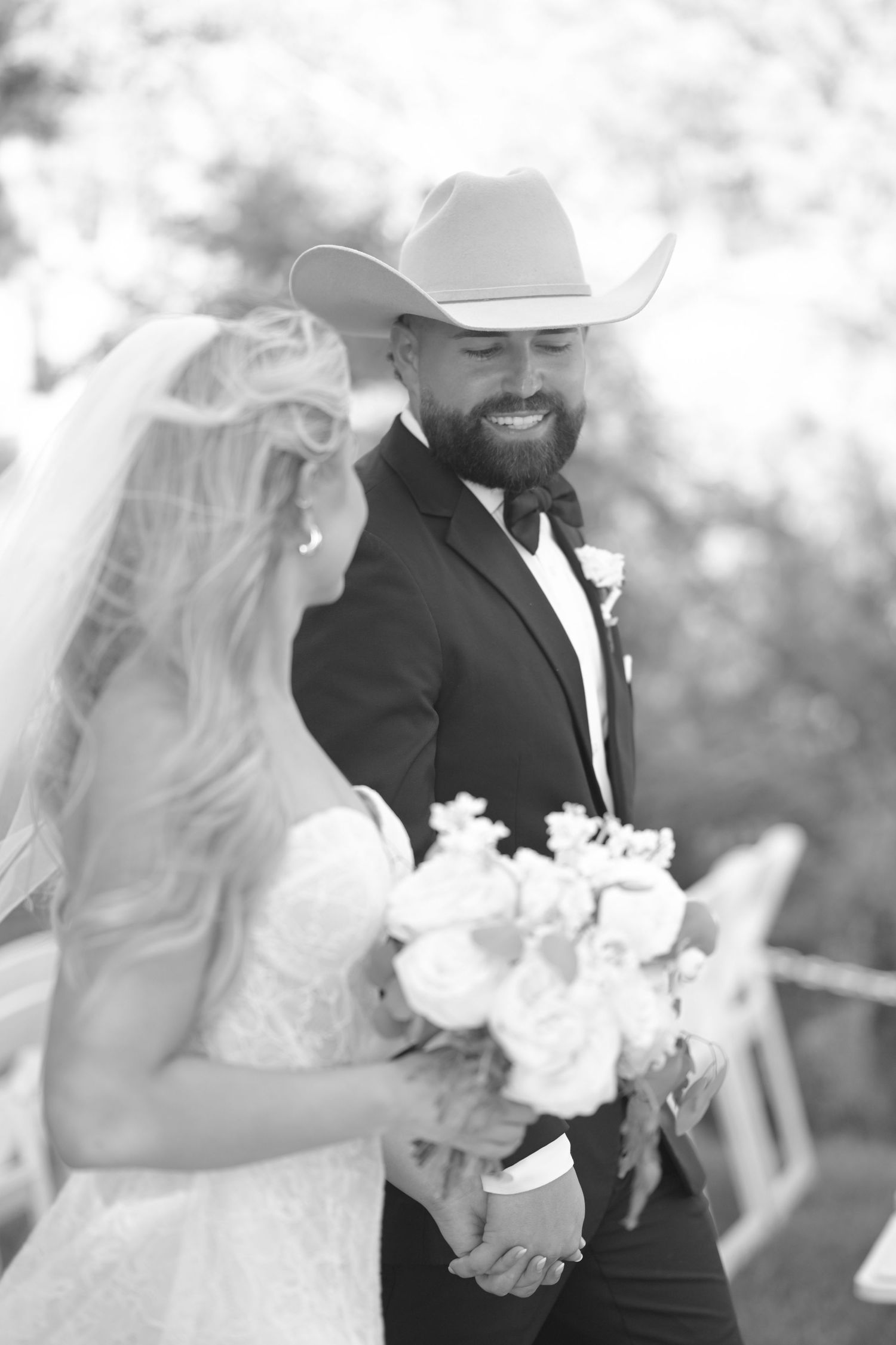 Black and white photo of smiling bearded groom in cowboy hat holding hands with bride holding bouquet.