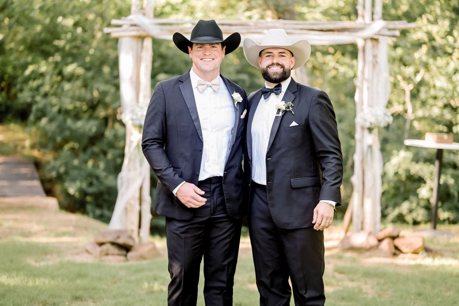 Two men in black suits and cowboy hats pose together in front of a rustic wooden arch outdoors.