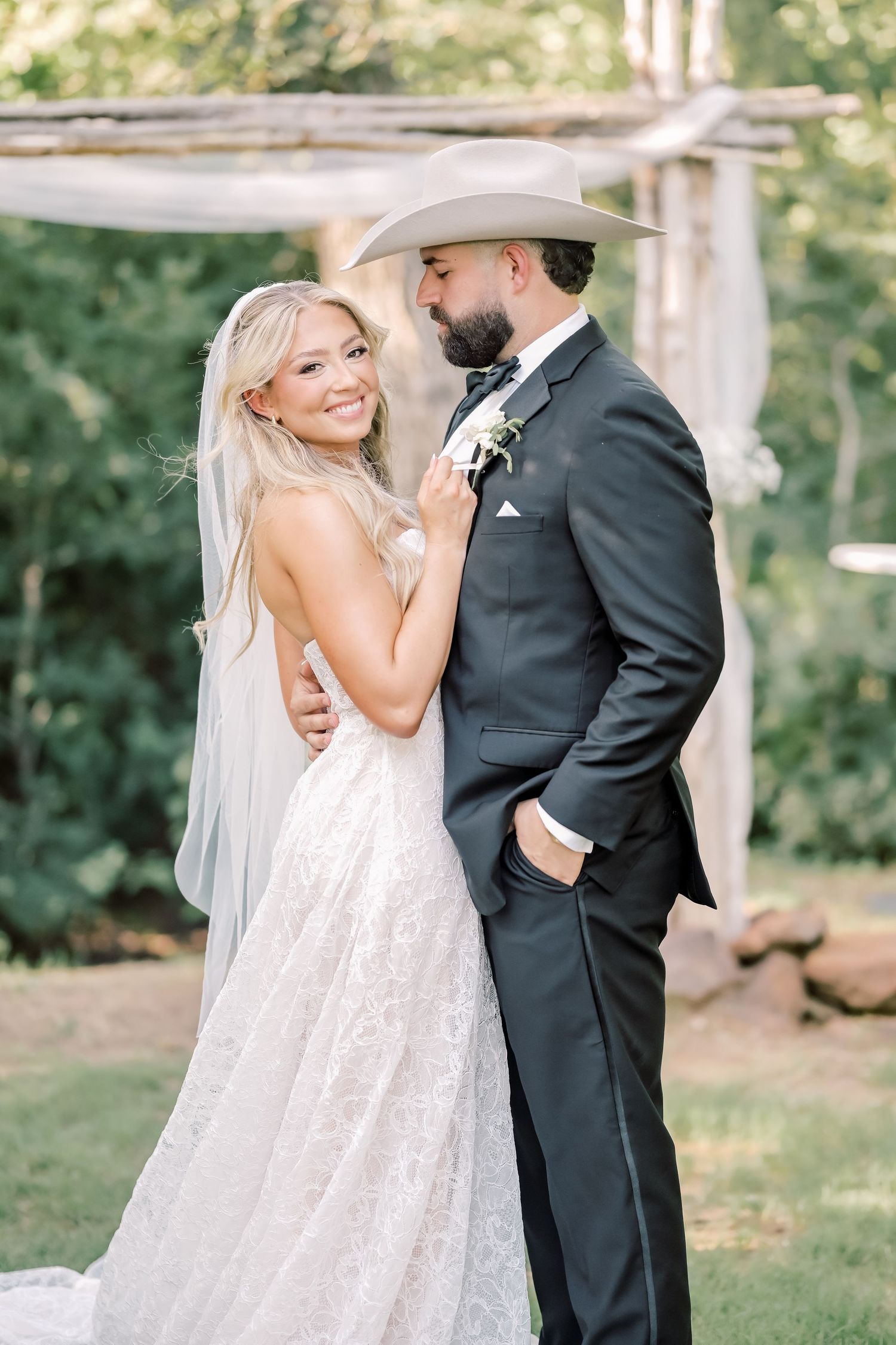 Bride in lace gown with veil embracing groom in black suit and cowboy hat at outdoor ceremony.