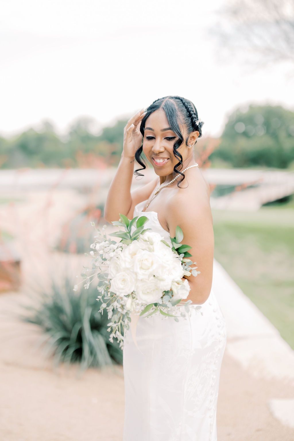 Smiling bride in white lace gown holds elegant bouquet of white roses and greenery outdoors.