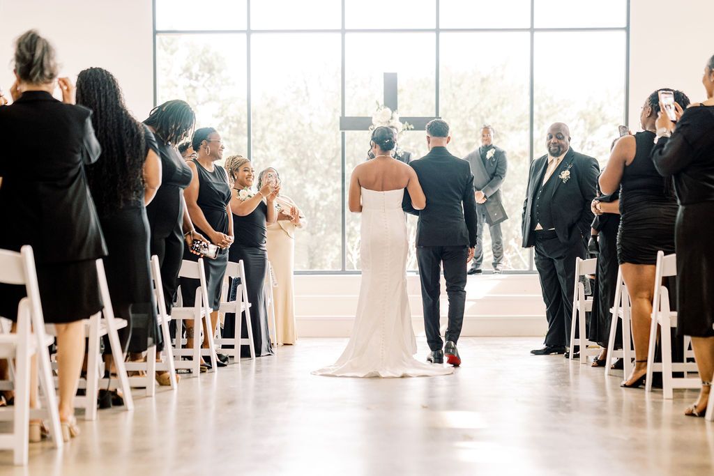 Bride and escort walk down aisle at modern indoor wedding ceremony with guests seated in white chairs.