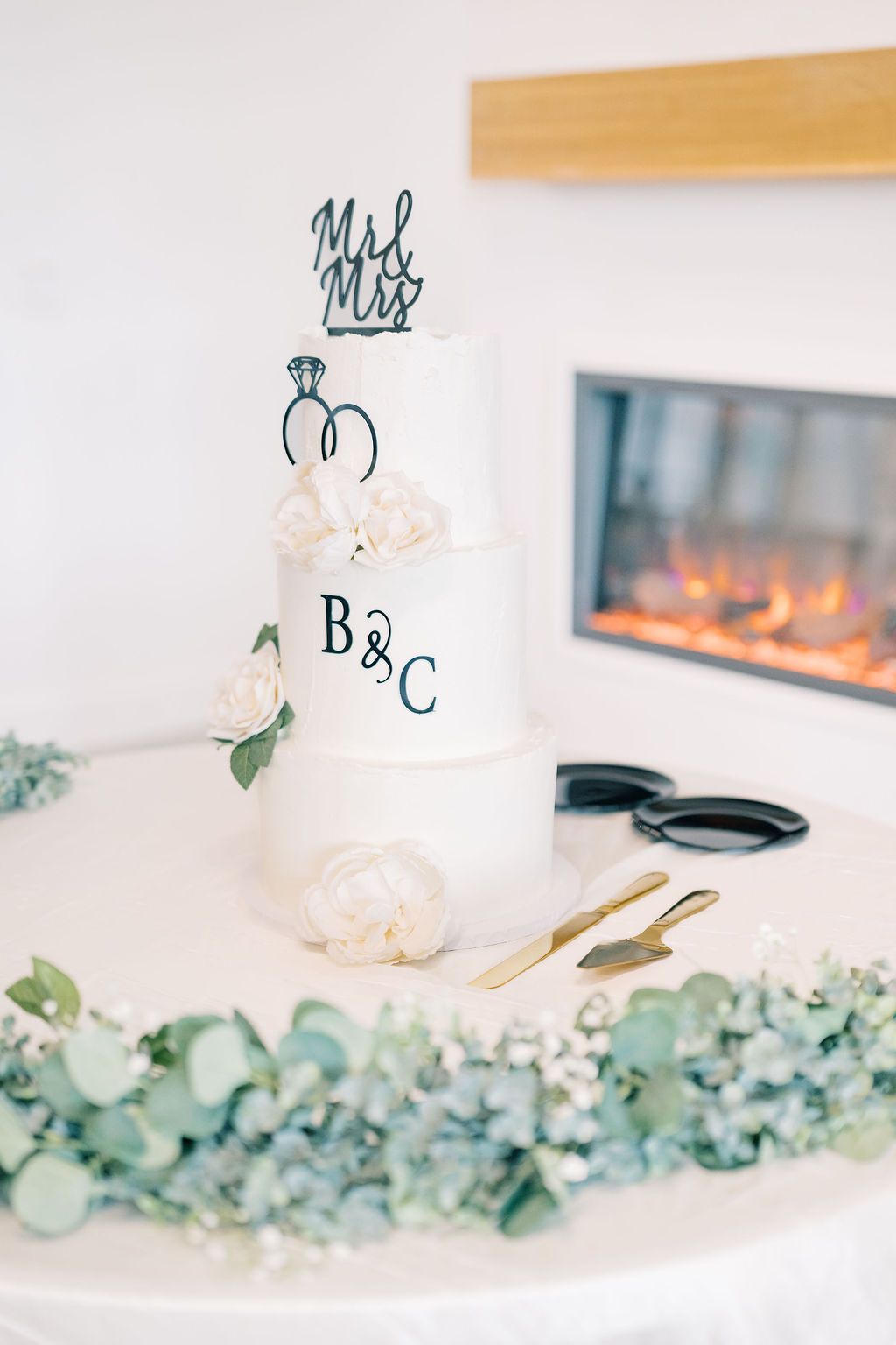 Elegant three-tier white wedding cake with Mr & Mrs topper, monogram B&C, floral decor, and gold serving set by a fireplace.