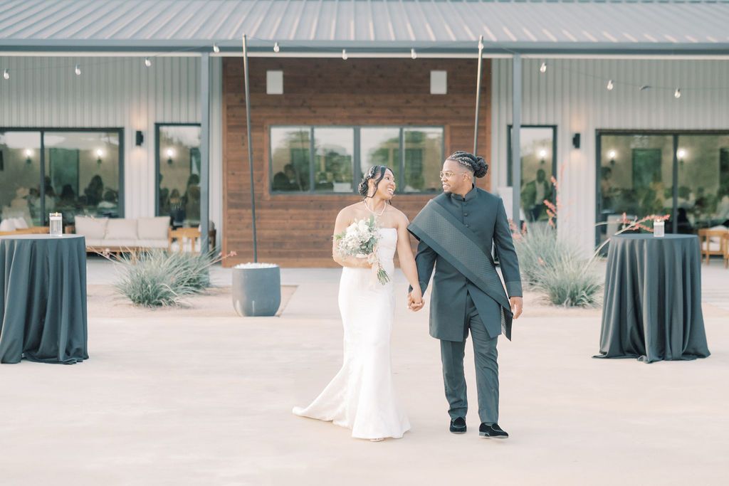 A bride and groom hold hands outside a modern wedding venue with string lights and elegant outdoor decor.