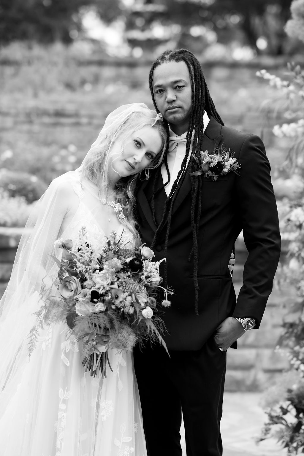 Black and white portrait of an interracial bride and groom posing with a floral bouquet at an outdoor wedding.