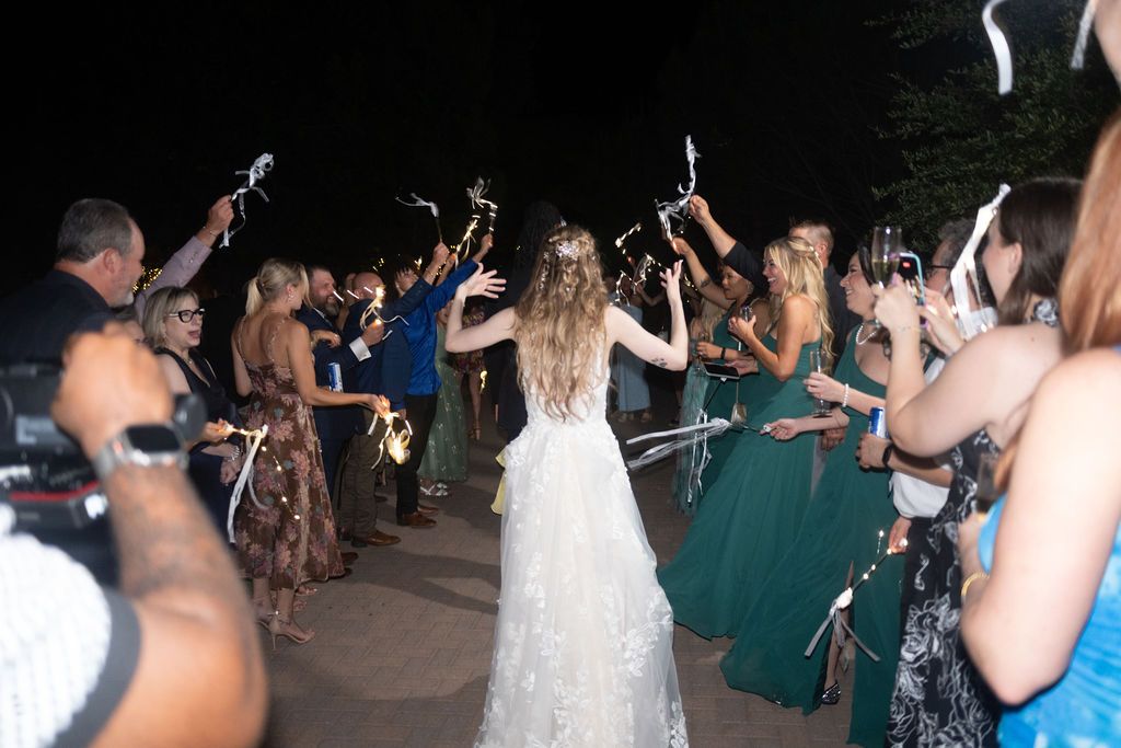 Bride in lace gown walks through cheering guests waving streamers at outdoor nighttime wedding celebration.