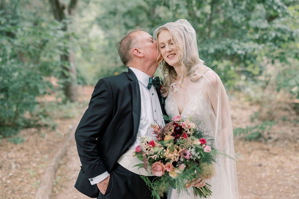 Groom in black tuxedo kissing bride's cheek as she smiles holding a lush floral bouquet in a wooded outdoor setting.