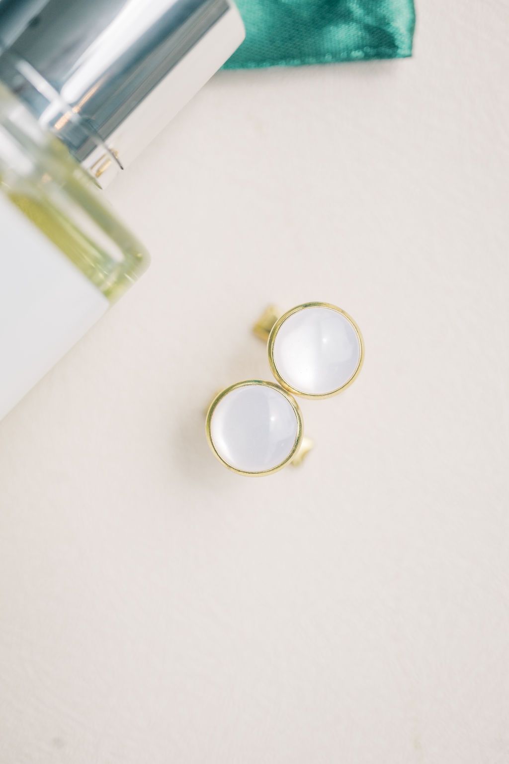 Gold compact mirror open on a beige surface beside a glass perfume bottle and teal ribbon.