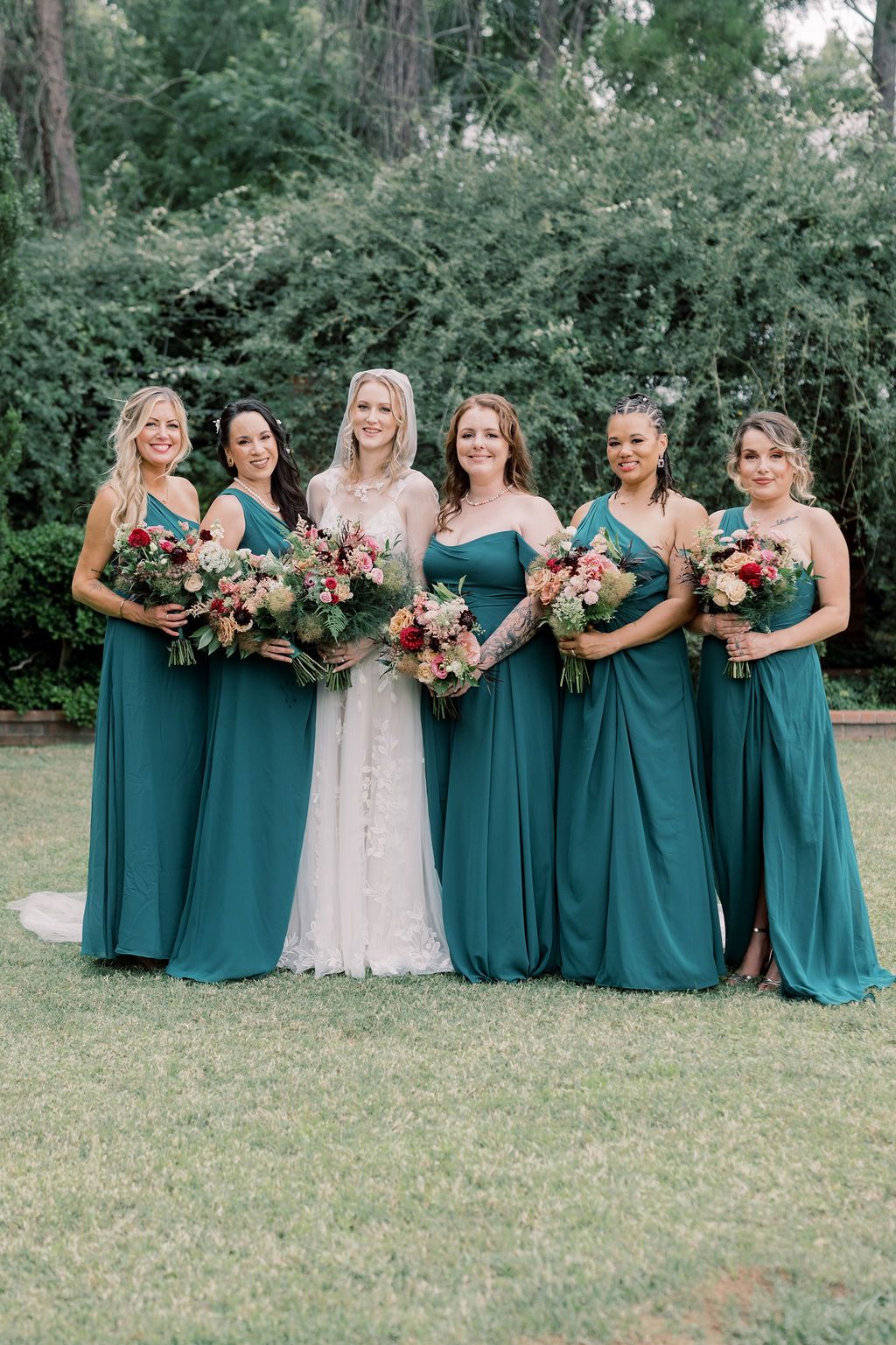 Bride in white lace gown poses with five bridesmaids in teal dresses holding colorful bouquets outdoors.