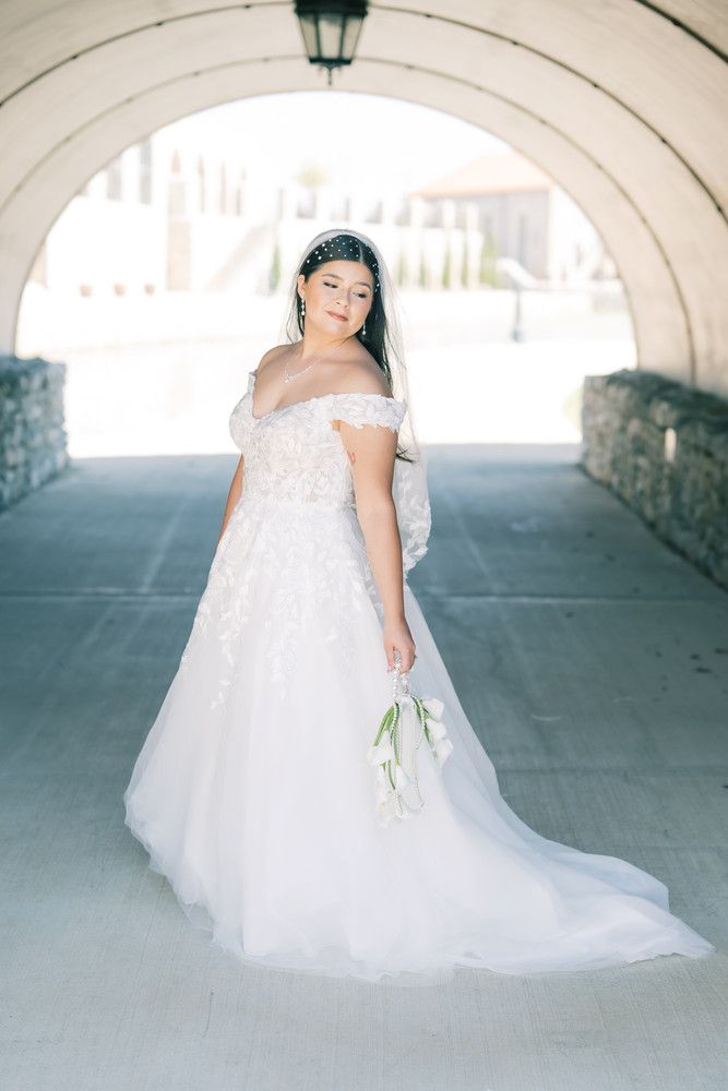 A bride in an off-shoulder white wedding gown poses elegantly under a stone archway.