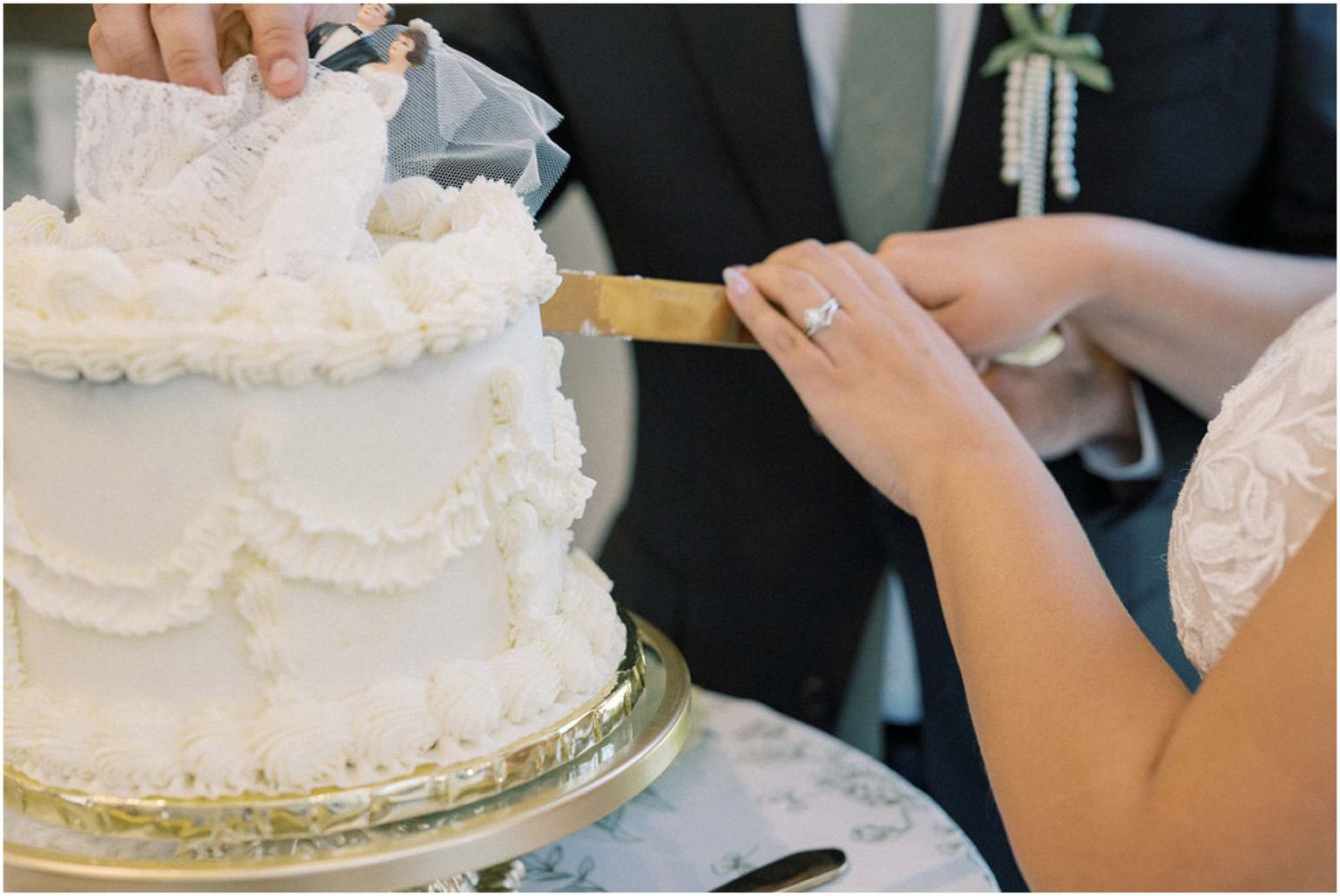 Hands cutting into a white tiered wedding cake decorated with delicate lace patterns and pearls.