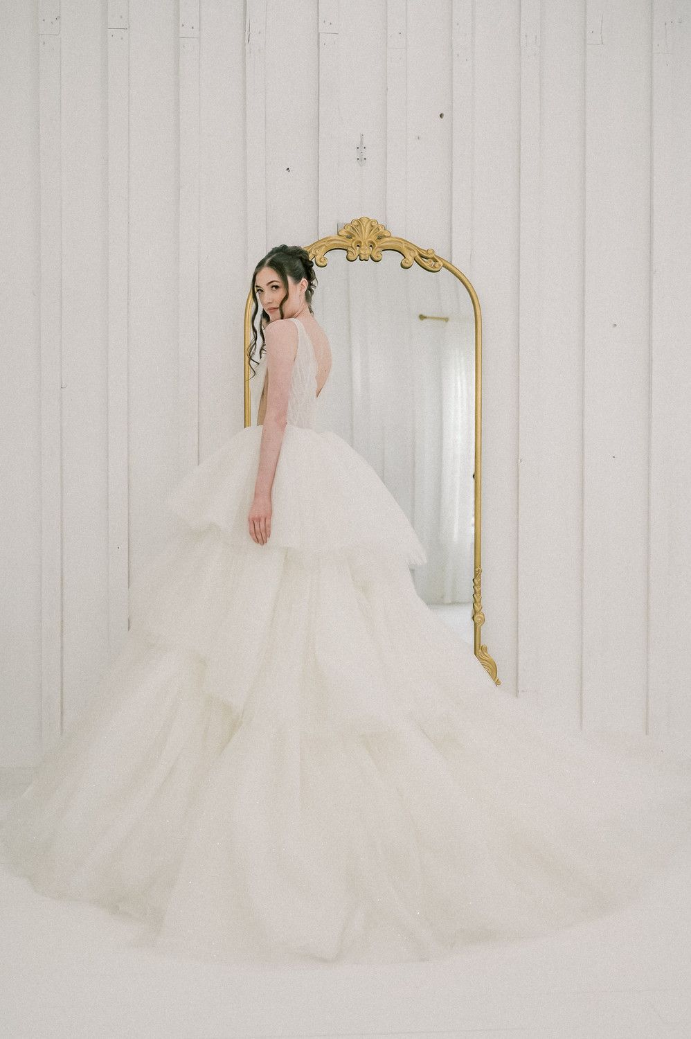Elegant wedding dress with voluminous tulle skirt reflected in ornate gold-framed mirror against white paneled wall.