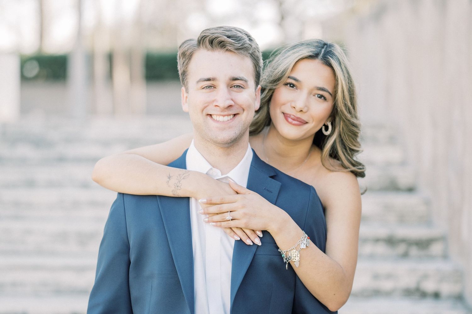 A couple poses together outdoors for an engagement photo, with the woman embracing from behind in a romantic moment.