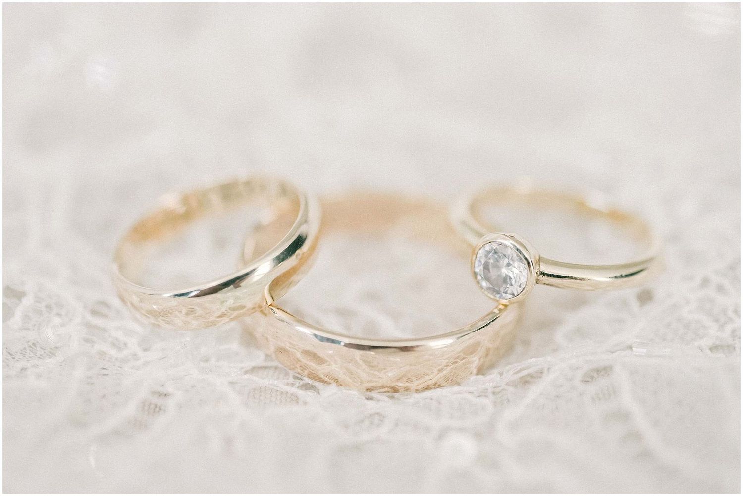 Three wedding rings including two gold bands and one diamond engagement ring arranged on delicate white lace.