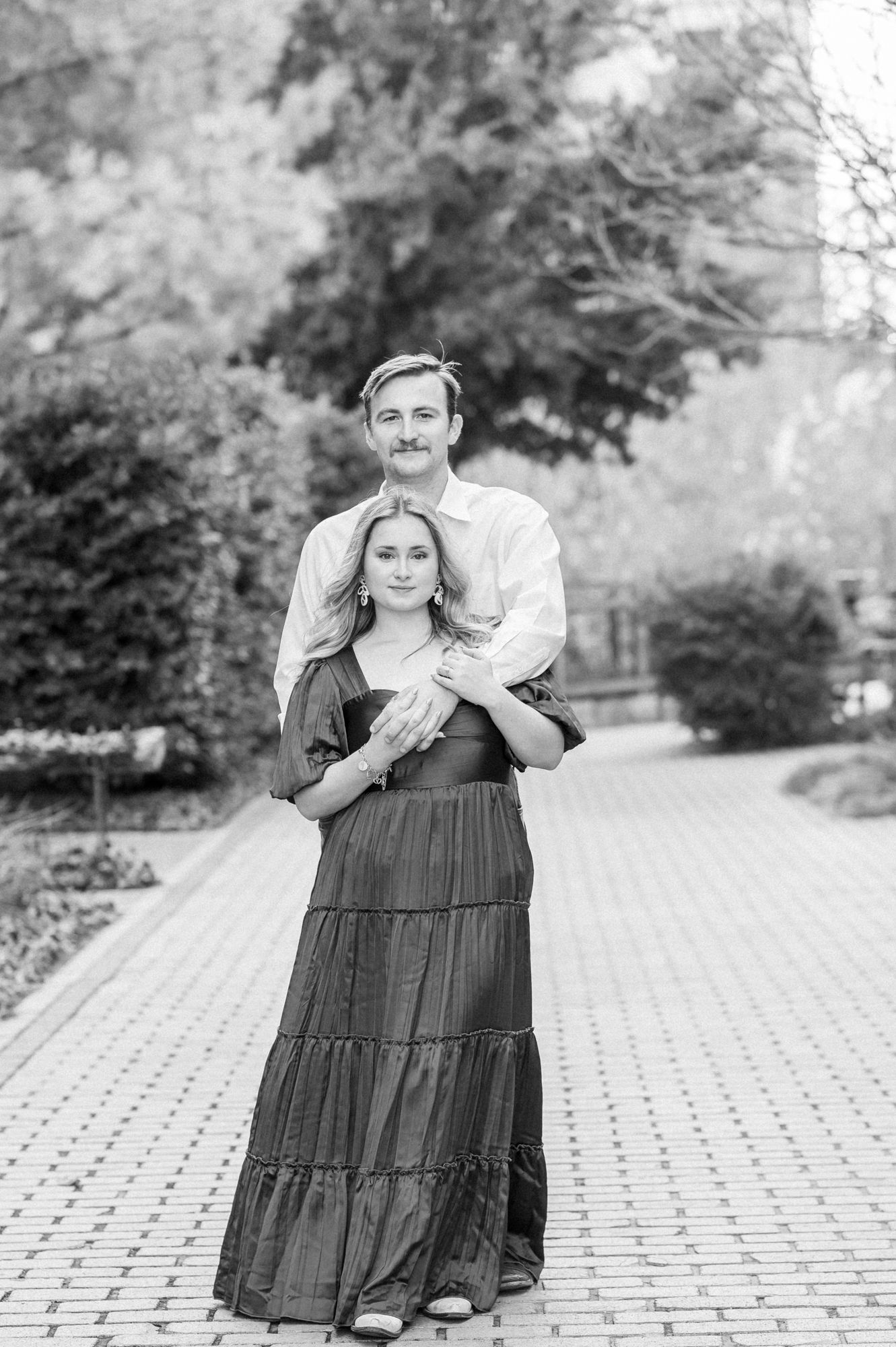 A couple shares a tender moment while posing together on a paved outdoor walkway surrounded by greenery.