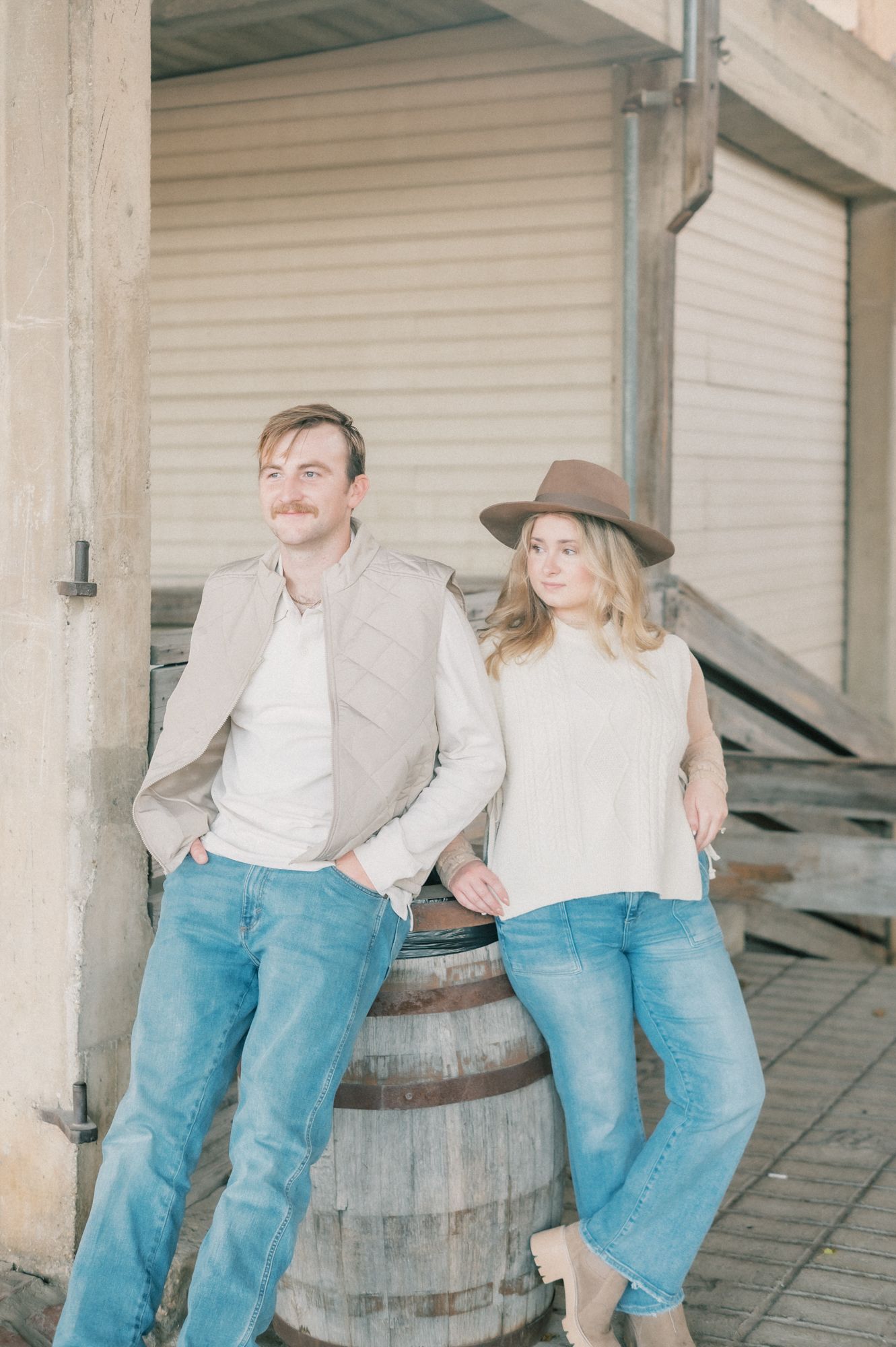 A couple in casual western attire poses playfully near wooden barrels and white garage doors.