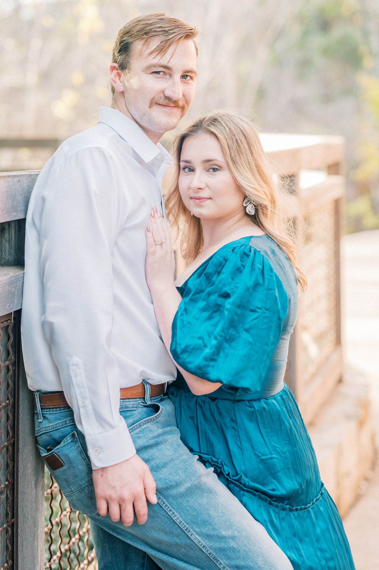 Couple embraces on rustic bridge with woman wearing teal dress and man in casual white shirt and jeans.