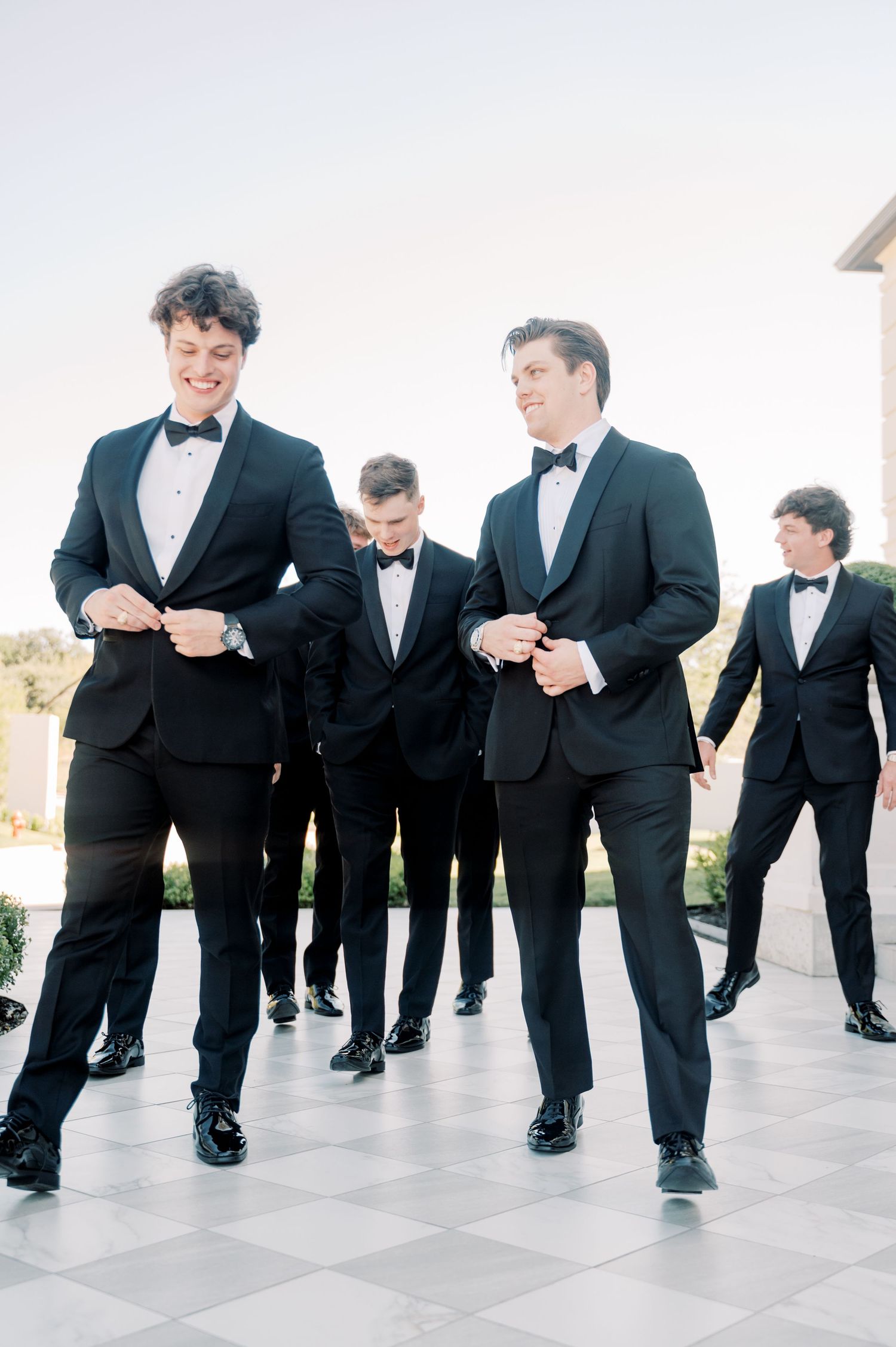 Groomsmen in black tuxedos walk in line while adjusting their bow ties and suits.