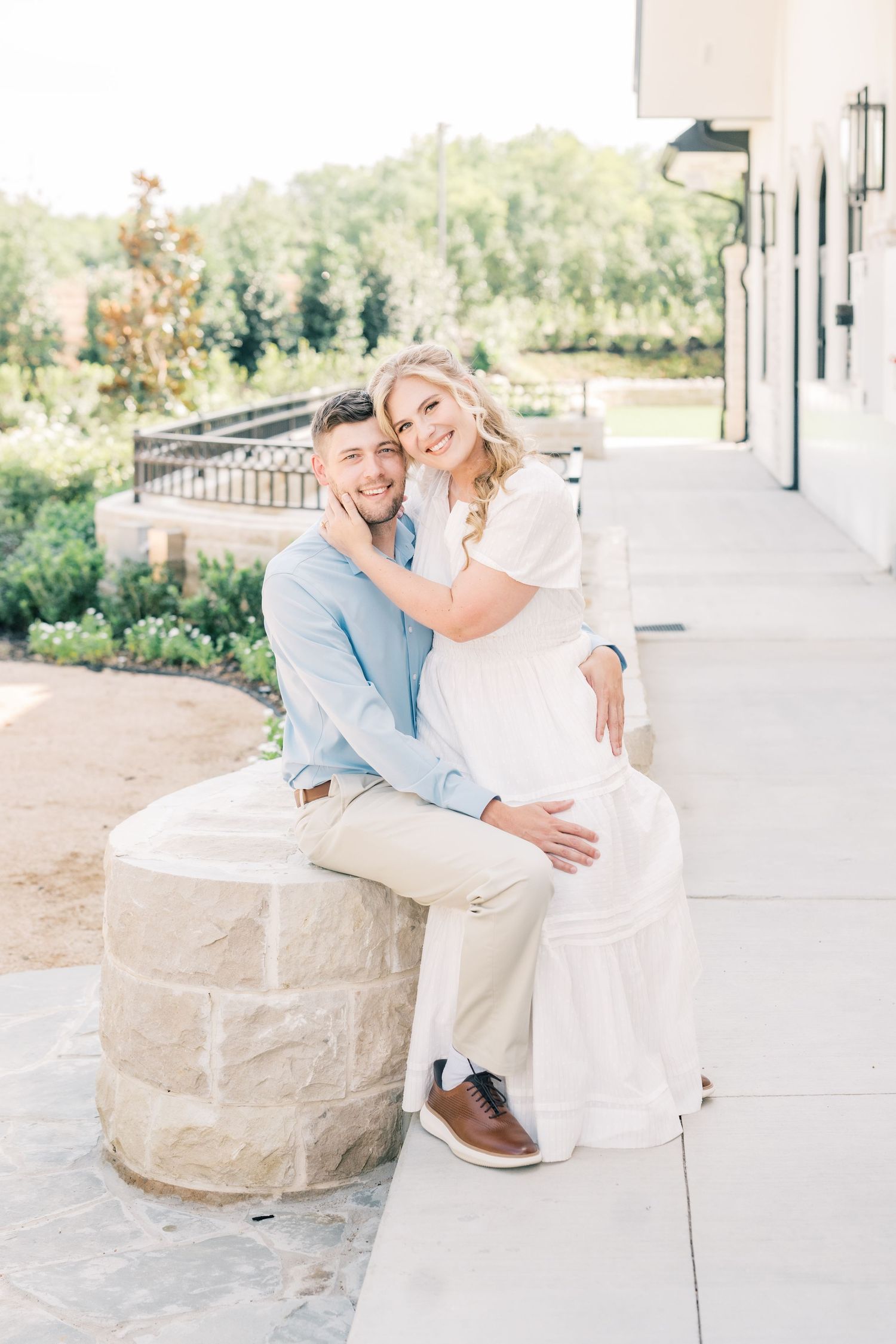 A happy couple poses outdoors, the woman in white embracing the seated man in a light blue shirt.